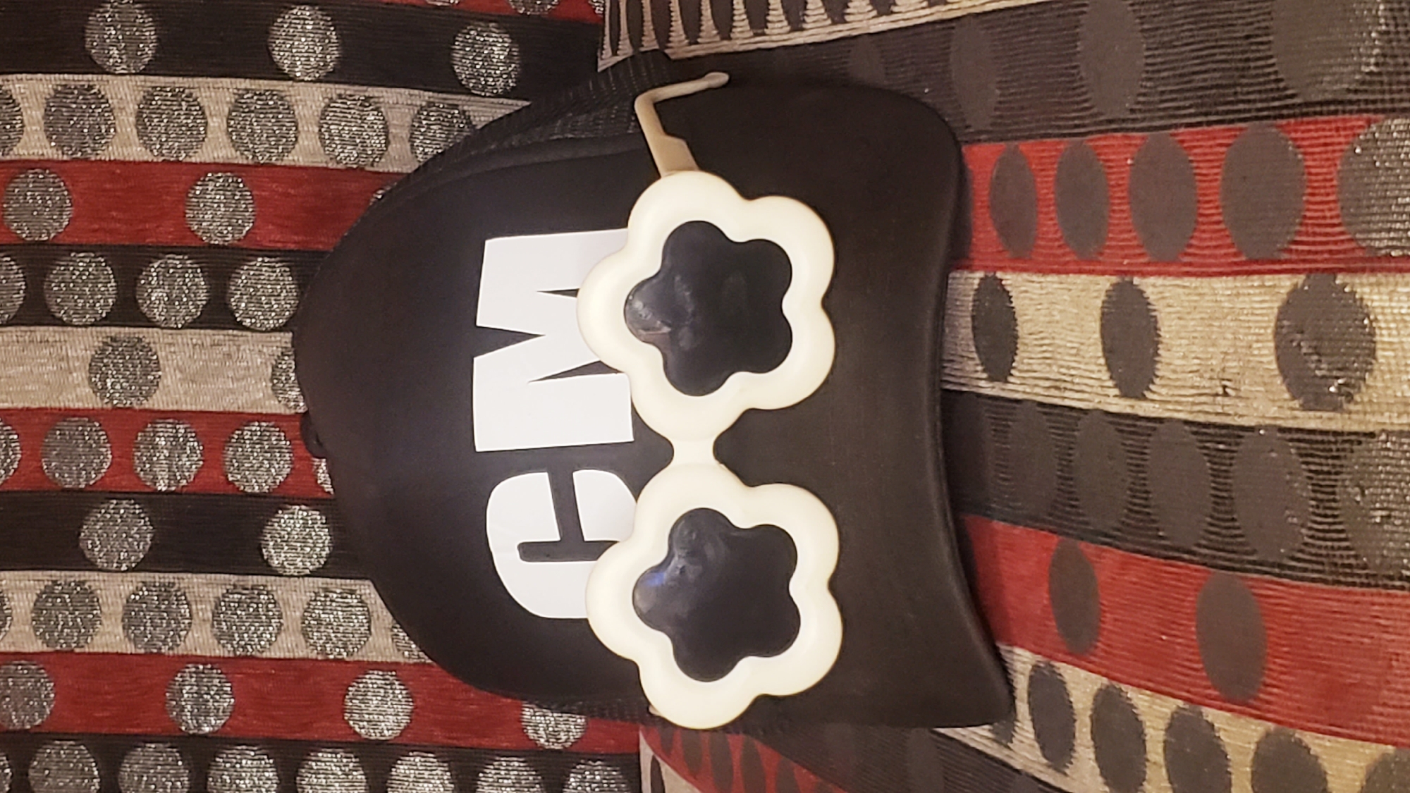 A black swim cap with the word "SWIM" in white block letters is lying on a patterned fabric. The fabric has horizontal stripes of red, black, and metallic silver, each with a row of dots. A pair of white, flower-shaped sunglasses sits atop the swim cap. The scene appears to be indoors. The location is not discernible as Narayi, Nigeria based solely on the image content.