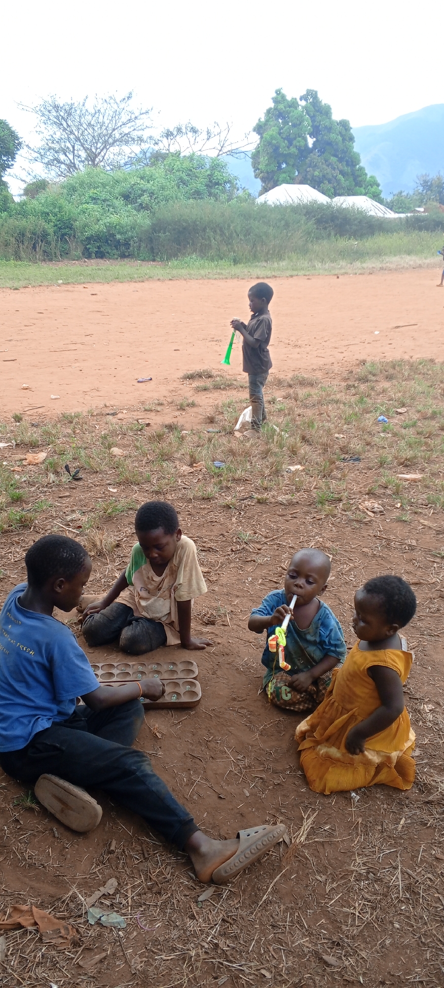 In an outdoor setting in Ngalula, Democratic Republic of the Congo, a group of children are engaged in various activities. Two boys are playing a traditional board game with holes and marbles, while a younger child is blowing a colorful plastic toy. A small girl in a yellow dress sits beside them, watching. Another child stands in the background holding a green plastic toy. The environment consists of a dirt ground with scattered grass and trash. Trees and a hazy mountainous landscape are visible in the background.