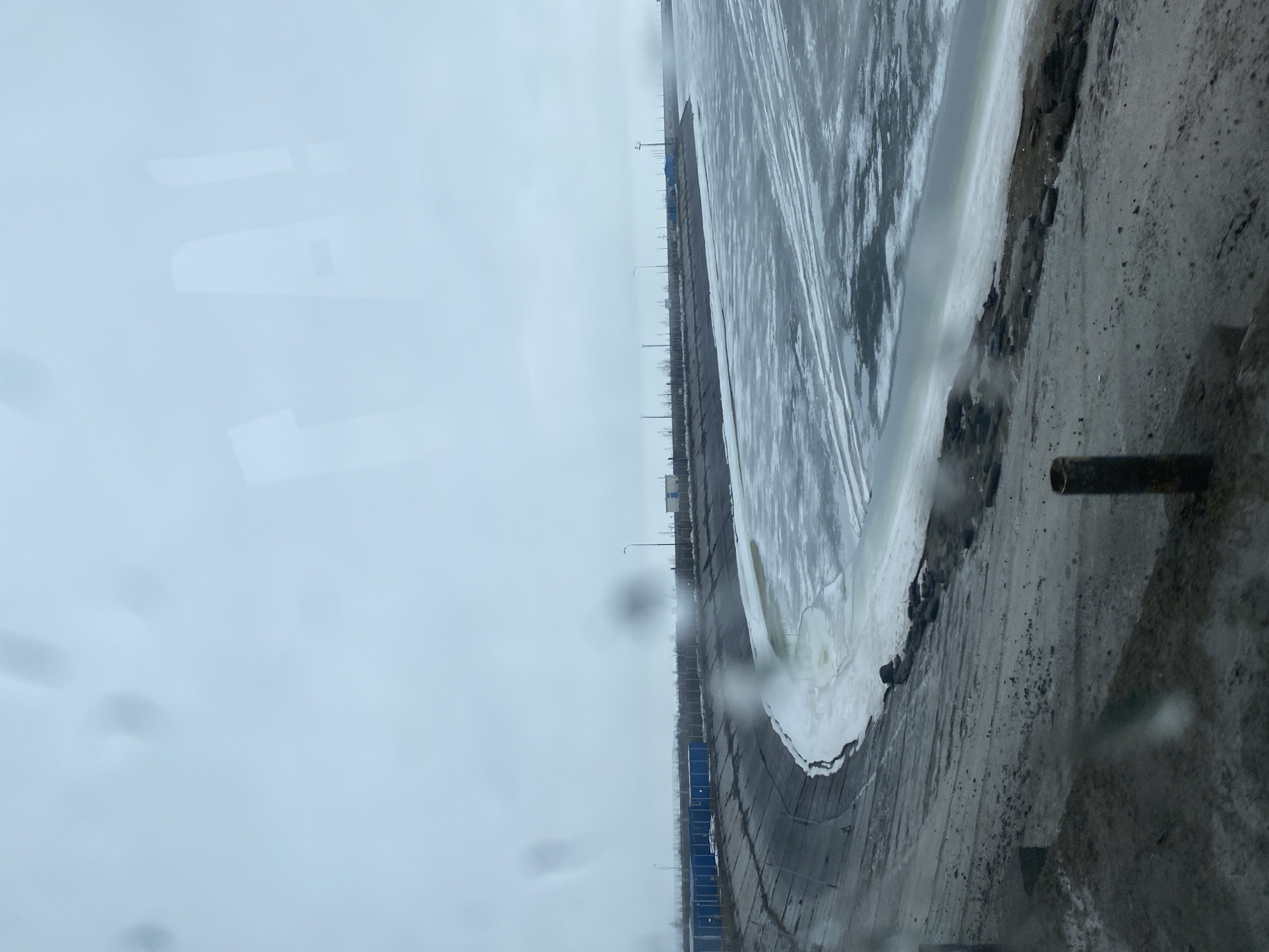 The image shows a view of a body of water, partially covered with ice, next to a concrete or paved area. There is a line of buildings or structures visible in the distance along the water. The overall scene appears overcast. There is condensation on the glass, indicating that it was taken from inside a window. The photo was taken in the city of Chaikovsky, Russia. The composition suggests an industrial or urban waterfront setting.