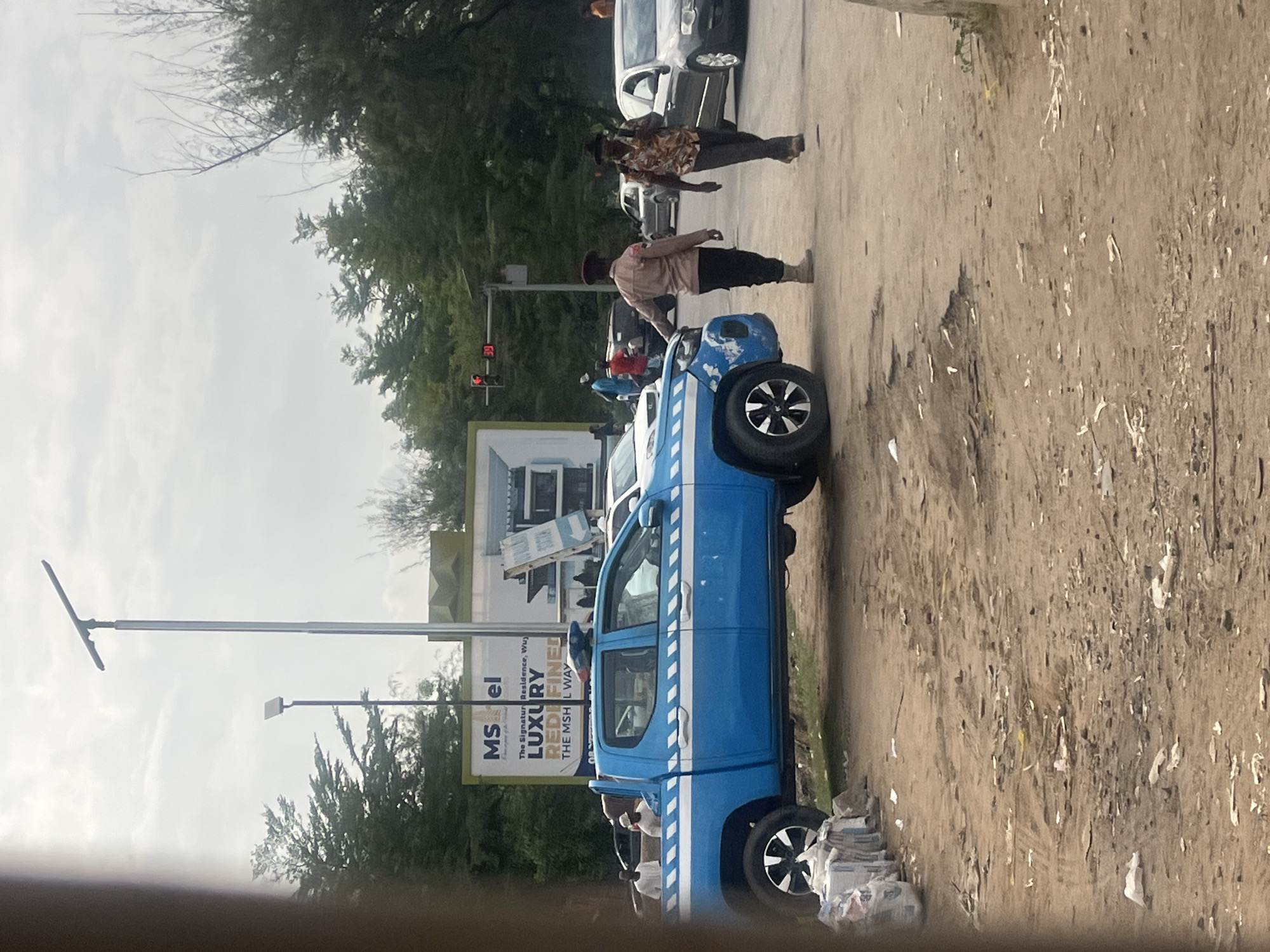 The image captures a daytime street scene in Karum, Nigeria, under an overcast or hazy sky. A dusty, unpaved road dominates the foreground and extends into the background, flanked by trees and foliage. Litter, including scattered papers and debris, is visible on the ground.
In the immediate foreground, a bright blue pickup truck, possibly a Toyota Hilux, is prominently positioned on the right side of the road. It features distinctive white diagonal stripes along its side, suggesting it might be a utility or official vehicle. Piles of newspapers or paper waste are stacked on the ground near its front wheel.
Several people are present in the scene. One individual, dressed in a light brown or beige shirt, dark pants, and a dark hat, stands near the front of the blue truck, appearing to be an officer or security personnel. Further down the road, another person in a patterned shirt and dark pants, also wearing a dark hat, walks away from the camera. Other less distinct figures are also visible.
Vehicles include a dark-colored car or SUV partially obscured in the mid-ground, and a lighter-colored car further back. Infrastructure elements include a tall metal street light pole on the left side of the road, topped with a flat, rectangular light fixture. A traffic light showing red is visible in the background, with a digital countdown display showing "39" (possibly seconds remaining) and "08". Behind the blue truck, a large billboard reads: "MSel", "The Signature Residence, Wu)", "LUXURY", "REDEFINED", and "THE MSH IL WAY".
The scene depicts a moment of everyday life, possibly at an intersection where traffic is stopped due to the red light. The overall atmosphere is mundane and observational.