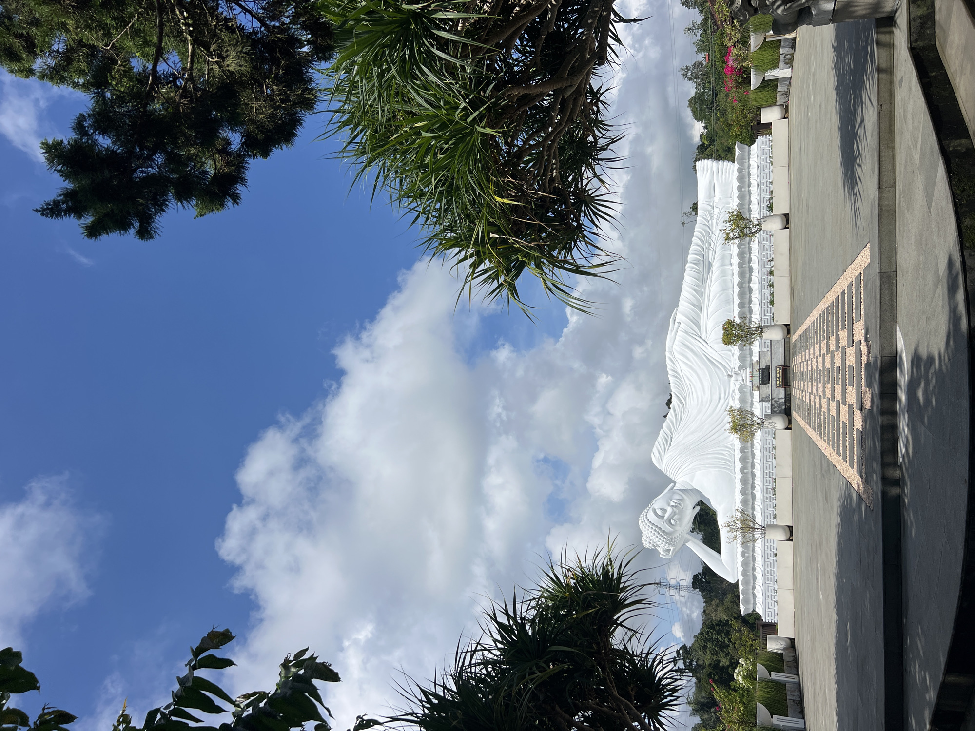 The image depicts a large, white reclining Buddha statue at Brahmavihara-Arama, a Buddhist monastery in Pupuan, Indonesia. The statue rests on a platform with decorative edging and small potted trees. A paved walkway extends from the base of the statue. The sky is visible above, with scattered clouds and blue sky. Trees frame the upper and lower edges of the frame, with foliage partially obscuring the view.