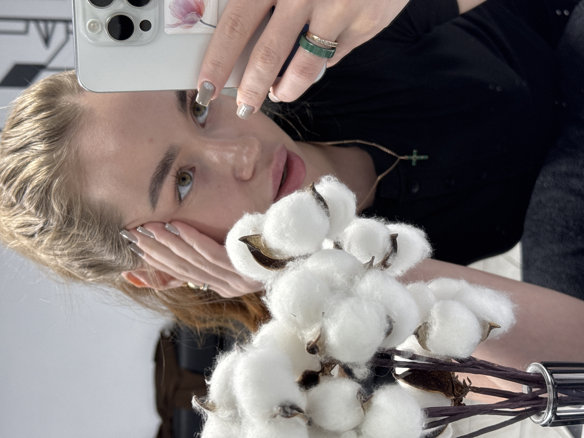A young woman takes a selfie with an iPhone 13 Pro or newer model. She has light blond hair, green eyes, and manicured fingernails. She's wearing rings on her hand holding the phone and a necklace with a green cross pendant. A bouquet of cotton flowers is visible in the foreground. The scene is likely indoors. The location of Chaikovsky, Russia is not evident from the image.