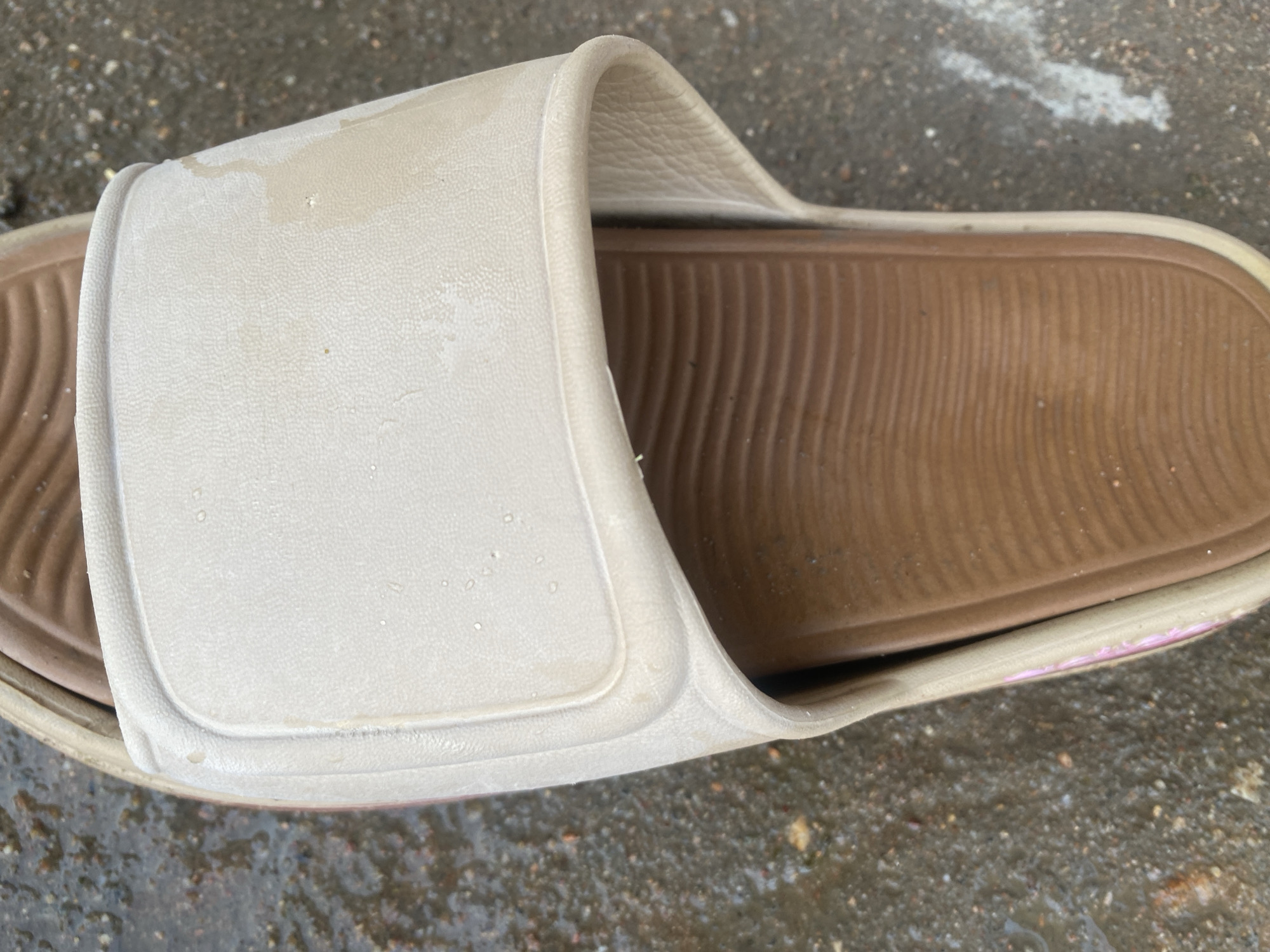 A close-up, overhead shot shows a tan slide sandal on a wet, grey, textured surface that appears to be pavement or concrete. The sandal has a brown, ribbed footbed and a wide, flat tan strap across the top. The strap has a slightly textured surface with small indentations, and there are a few small water droplets on it. The surrounding ground is wet and speckled with small, dark, pebble-like fragments, suggesting it may be an outdoor environment. A faint pink design is visible on the side of the sandal, near the heel. There are no people visible in the image. The scene is static and appears to have been captured in Jalingo, Nigeria.