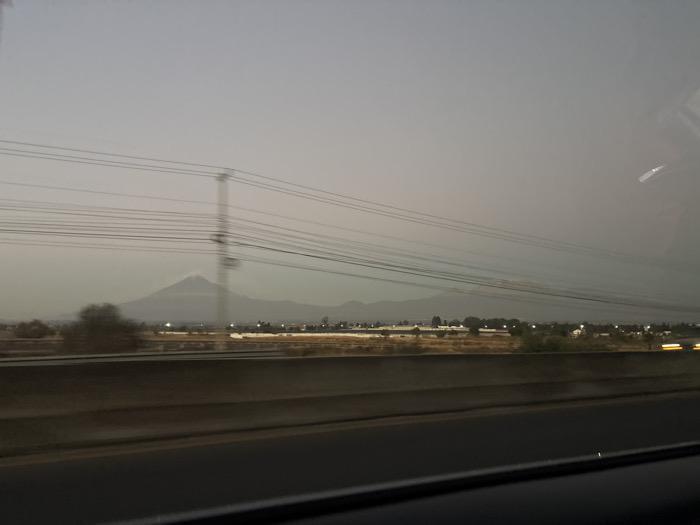 The image depicts an outdoor, hazy, twilight scene viewed from a vehicle on a highway near Santa María Zacatepec, Puebla, Mexico. In the immediate foreground, a dark vehicle interior frame borders the bottom. Above this, a dark concrete highway barrier spans horizontally. Beyond the barrier, a multi-lane asphalt road surface is visible. The midground features a flat expanse of dry, light-brown terrain with scattered low-rise buildings, possibly industrial or commercial, and sparse trees. Numerous dark overhead power lines run horizontally across the frame, supported by utility poles, including a prominent, vertically blurred pole near the center-left. Small, bright points of artificial light, some exhibiting motion streaks, are visible within the midground. The background is dominated by a large, hazy, conical mountain, identifiable as Popocatépetl, flanked by lower mountain ranges, all silhouetted against a uniform, muted gray sky. Reflections from a vehicle window are present in the upper right portion of the image. No individuals are discernible.