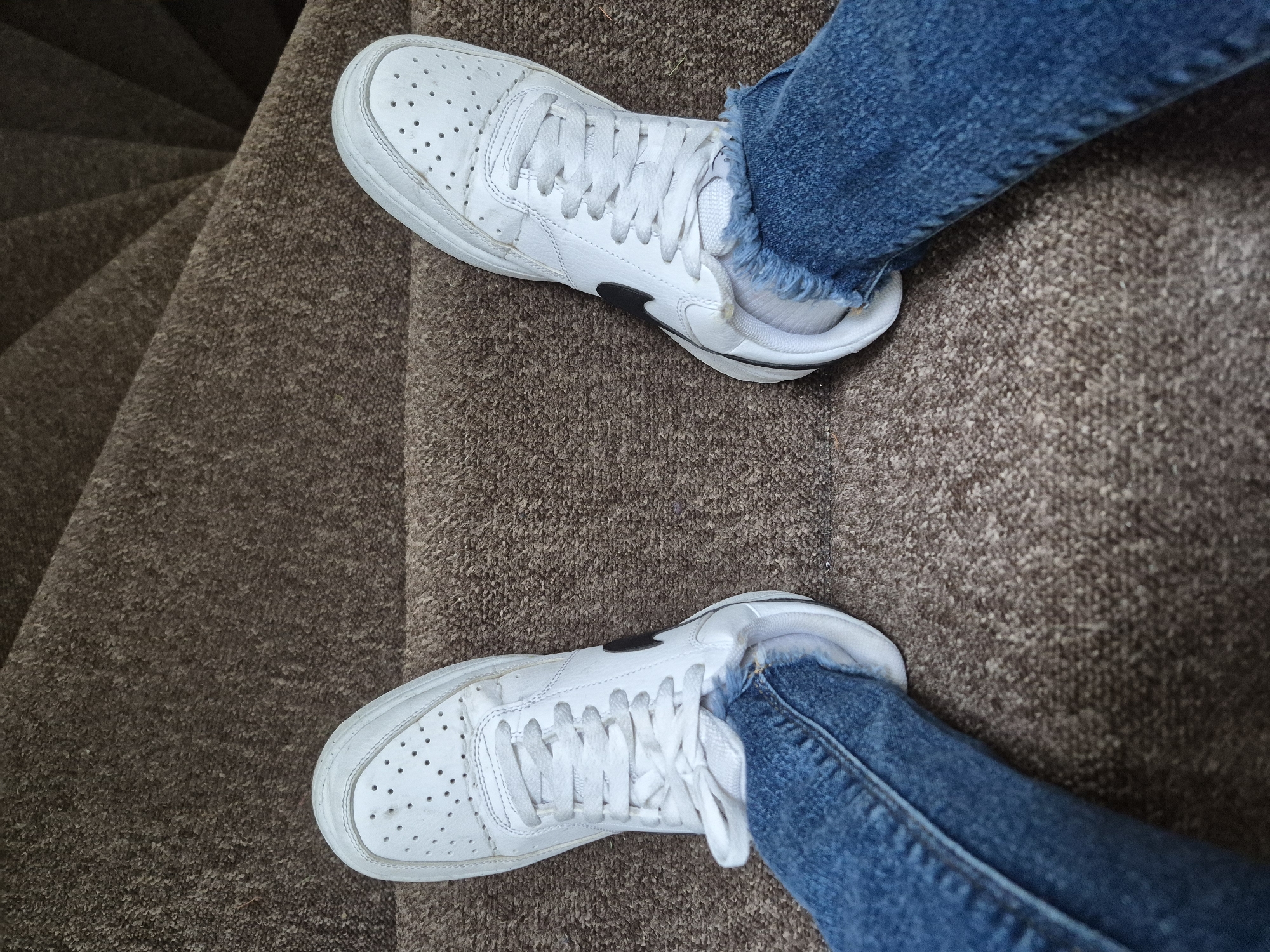 A first-person perspective shot shows a pair of white Nike Court Vision Low sneakers on a person's feet, who is wearing blue denim jeans. The sneakers have a black Nike swoosh logo on the sides. The person is standing on a textured brown carpeted surface, which appears to be stairs. The scene is lit by an overhead light source, creating subtle shadows. The image was taken in Sandwell, United Kingdom.