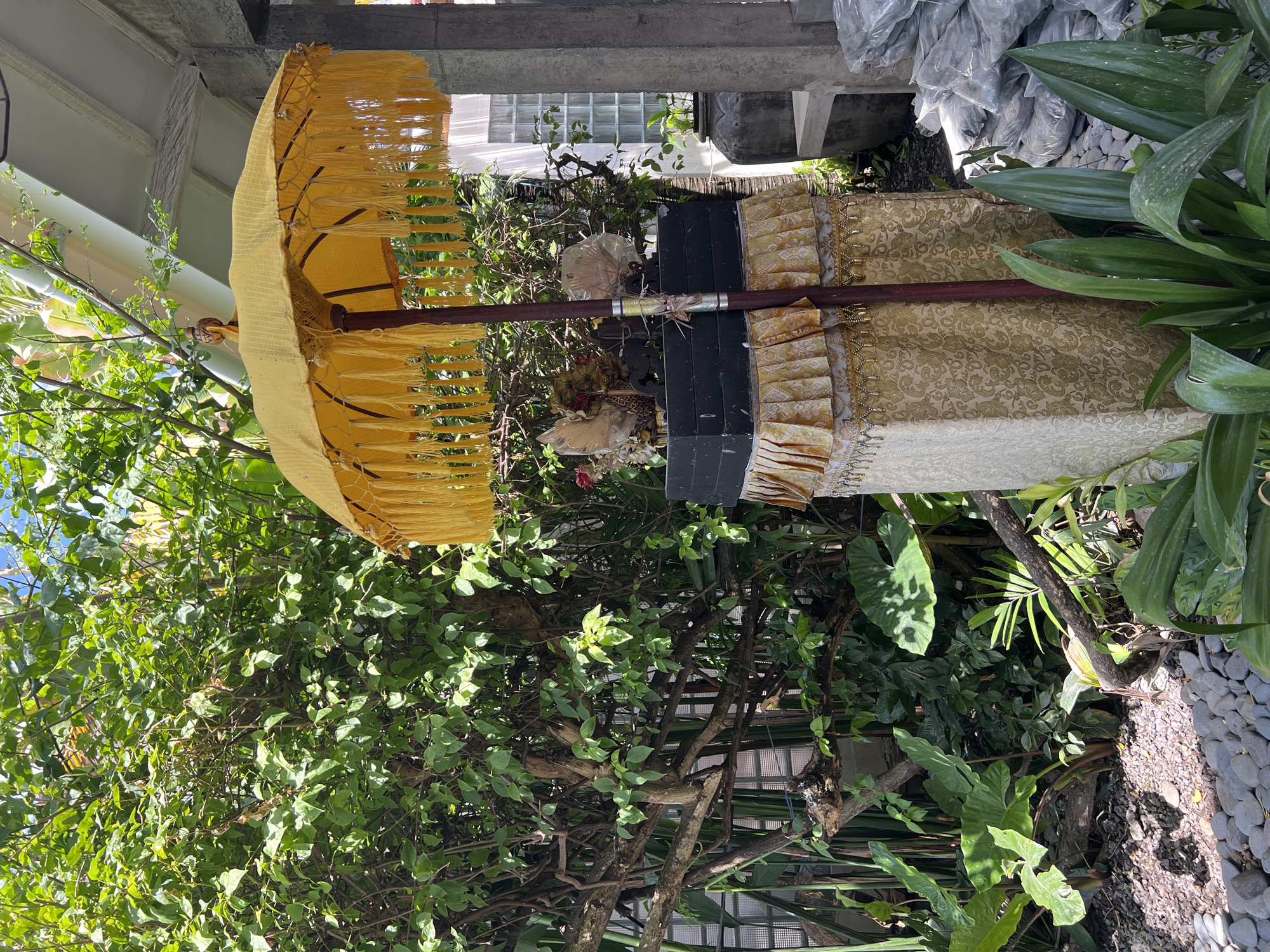 The image depicts a yellow Balinese ceremonial umbrella standing beside a floral offering, likely in a garden or outdoor area, possibly in North Kuta, Indonesia. The umbrella has a bamboo-like pole and a fringe of yellow tassels. The floral offering is placed on what appears to be a tiered, decorated stand. The surrounding environment includes various plants, shrubs, and some garden stones. A building structure is partially visible in the background.