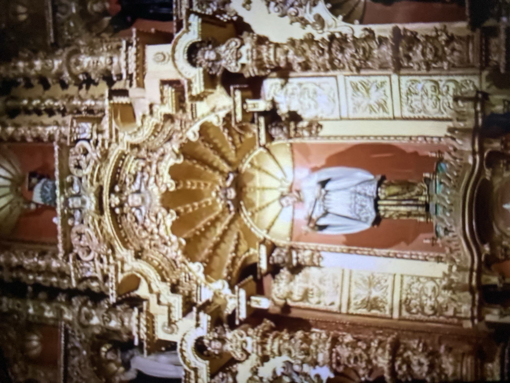The image depicts an elaborately decorated interior, likely a religious building, located in Tinaquillo, Venezuela. The scene is dominated by ornate gold-colored architectural elements, including intricate carvings and moldings. A statue, possibly of a religious figure, stands within a niche. The walls are painted red.