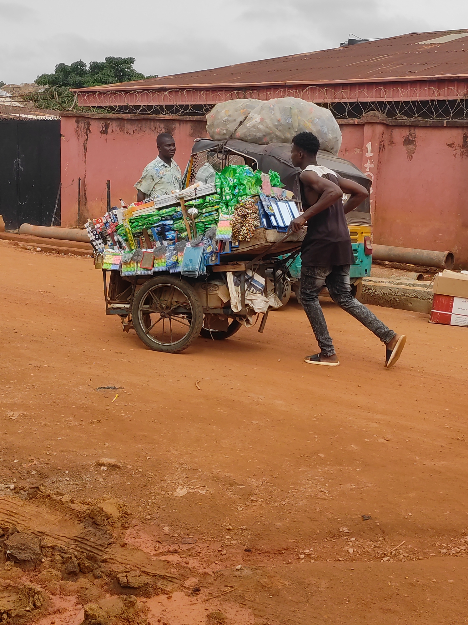 Two African men are present in the city of Jos, Nigeria. One man, wearing a short-sleeved shirt, stands behind a hand-drawn cart loaded with various items, including school supplies and snacks. The second man, wearing a tank top and jeans, pulls the cart forward. The cart is heavily loaded, including large bags filled with recyclable plastics. The ground is red earth. A building with a red roof and barbed wire along the top of the wall is in the background. A vehicle is near the cart in the background.