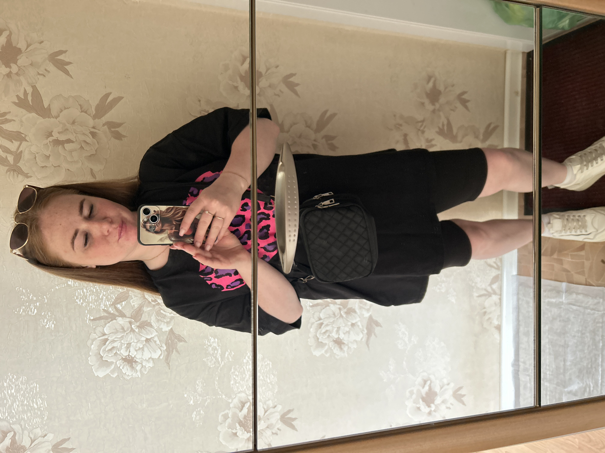 A young woman with long, reddish-brown hair and freckles is captured in a mirror selfie, holding a silver-colored smartphone with a decorative case. She wears sunglasses pushed up onto her head and has a relaxed expression with her eyes partially closed. Her attire consists of a black t-shirt or dress featuring a vibrant pink and purple leopard print design on the chest, along with a small black quilted fanny pack worn across her body. On her left wrist is a thin gold bracelet, and she wears two rings on her left hand. Her lower body is in black fabric, possibly shorts or a dress, exposing her bare legs, and she wears white sneakers.
The setting appears to be an indoor space within a residence in Chaikovsky, Russia. The wall behind her is adorned with light-colored wallpaper featuring a prominent, subtle floral pattern of large peonies or similar flowers in shades of cream and beige. The mirror itself appears to be a multi-panel reflective surface, and a silver-colored door handle is visible through the reflection near her midsection. The floor visible in the reflection includes both a dark red carpet on the right and a lighter, possibly wooden or tiled surface near her feet on the left. The activity taking place is a casual mirror selfie, likely indoors during the daytime. No legible text is visible other than the faint design on her phone case.