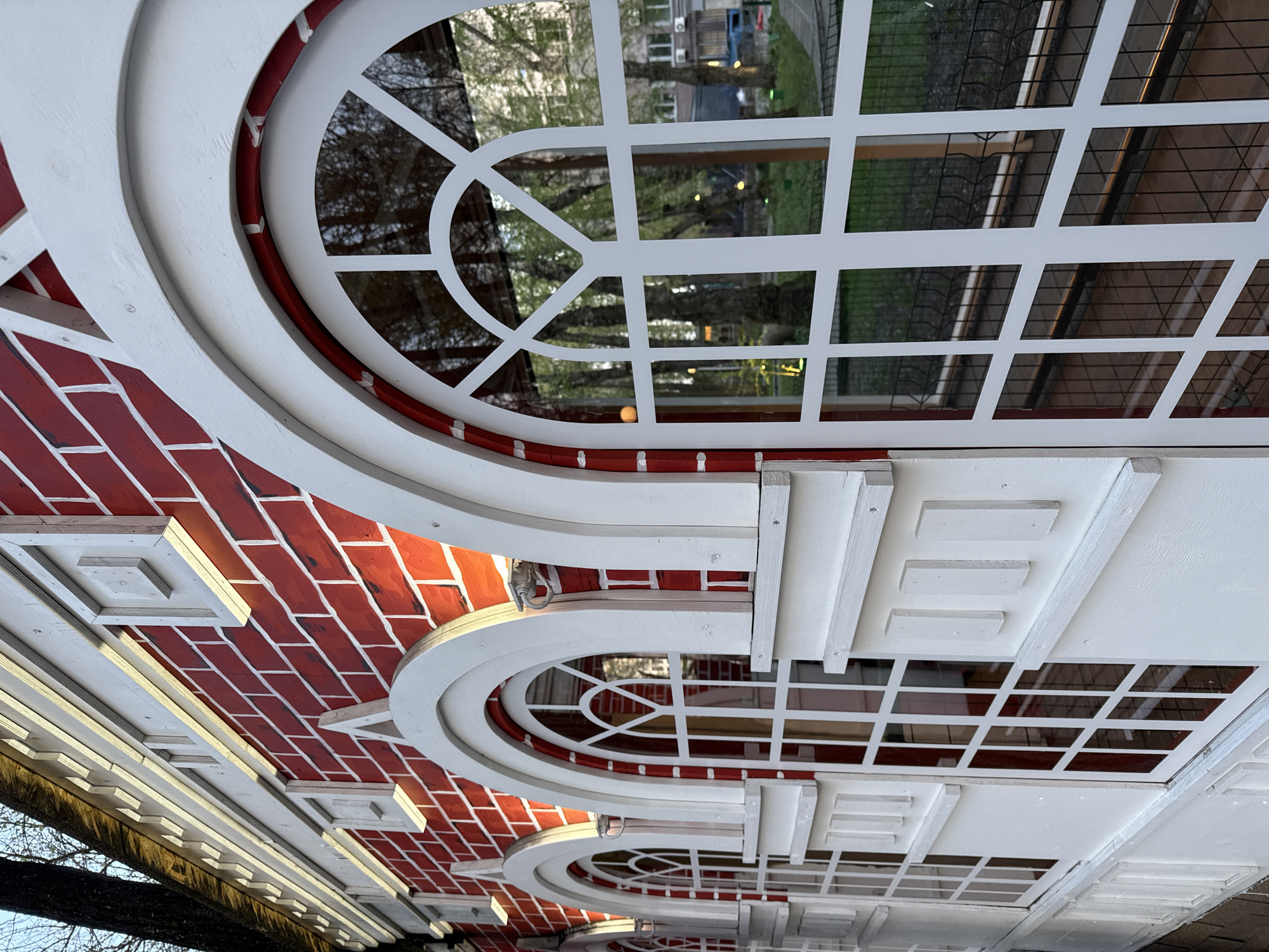 The image shows the facade of a building in Perm, Russia. The exterior features red brickwork, interspersed with decorative white window frames in an arched style. The windows have a complex grid pattern of white mullions and transoms. Reflections visible in the window panes suggest trees, buildings, and possibly street lighting, indicating an urban environment. The photograph appears to be taken at an upward angle, emphasizing the architectural detail of the building's exterior.