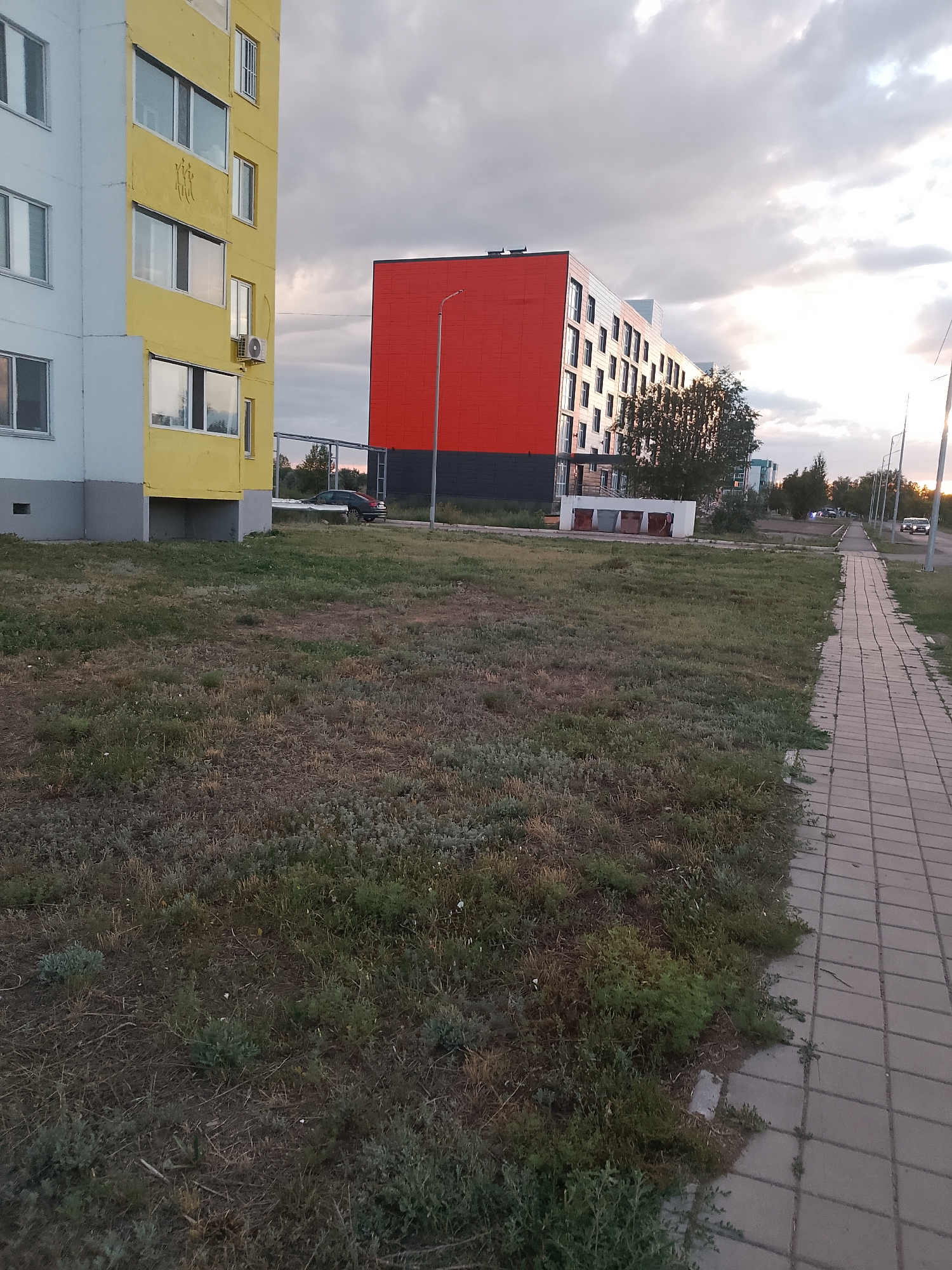 The image depicts a street scene in Abay, Kazakhstan. In the foreground, there is a patch of dry, overgrown grass. To the right, a paved sidewalk runs alongside the grass. Beyond the grass and sidewalk, there are several buildings. On the left, a yellow and white apartment building is visible, with multiple windows and an air conditioning unit. Further to the right, a modern building with a striking red facade and dark grey base stands prominently. This building has numerous windows and appears to be multi-story. In the background, more buildings, trees, streetlights, and a few vehicles are visible, suggesting a residential or mixed-use area. The sky is overcast with dramatic clouds, and the lighting suggests it is either early morning or late evening.