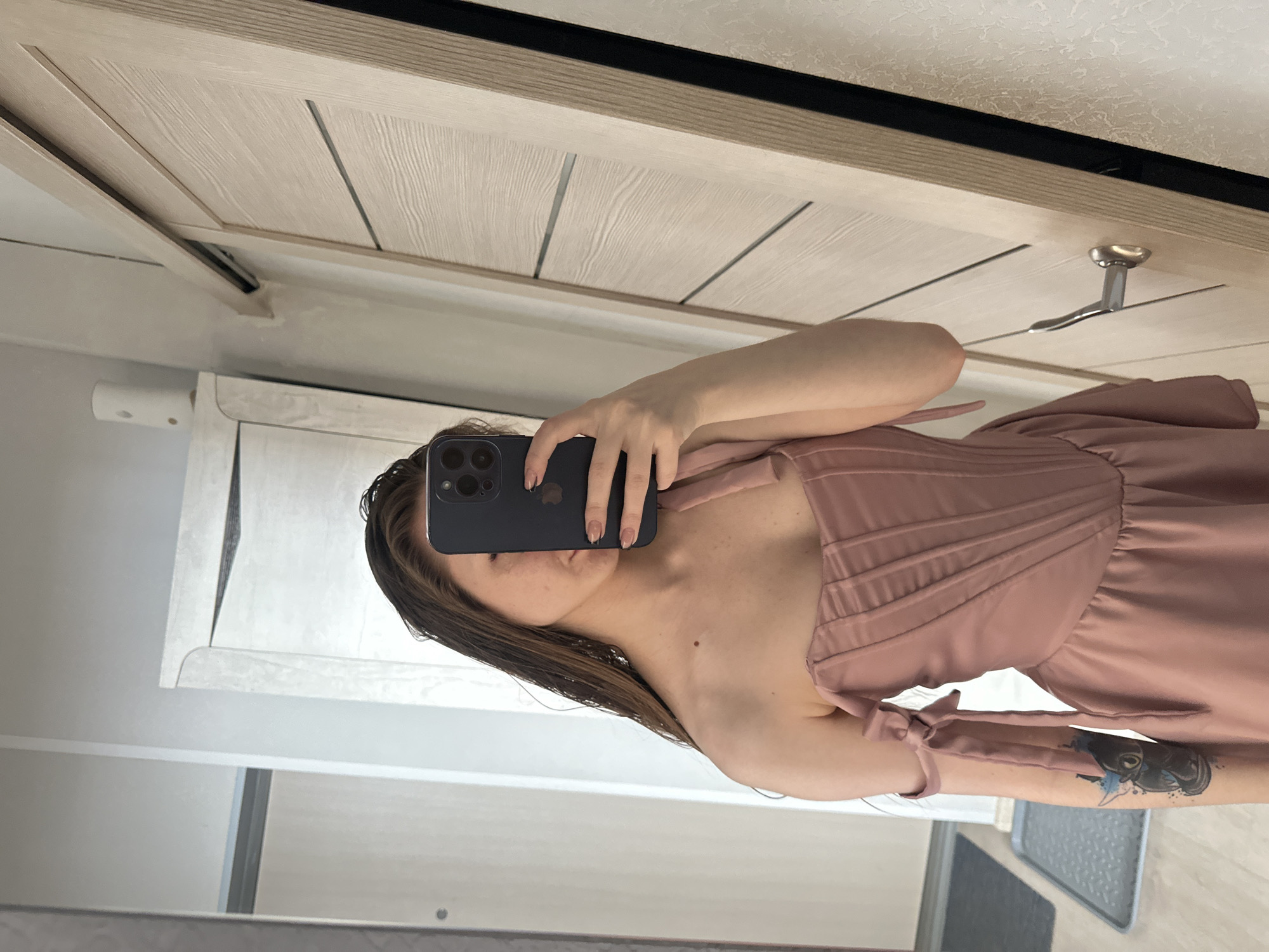 A woman is taking a selfie in a mirror. She is wearing a pink dress and holding a dark-colored smartphone in front of her face. Part of her upper body is visible. Her wet hair is dark. The reflection in the mirror shows a bathroom or dressing room setting with light-colored doors, a toilet paper holder, and a gray floor mat. The location appears to be indoors.
