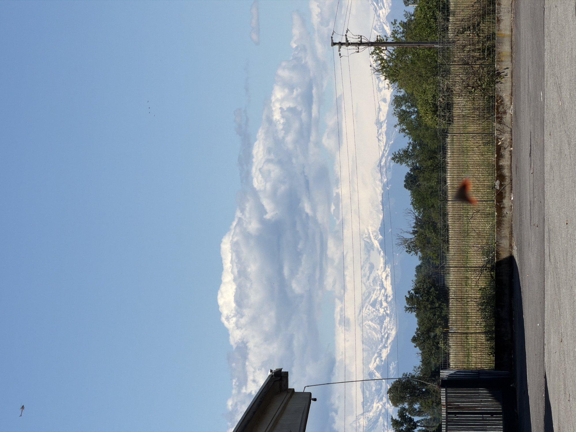 The image shows a street view in Gudo Visconti, Italy. On the right, there's a portion of a paved road, adjacent to a fence that borders a grassy field with trees. Power lines stretch across the frame, partially obscuring a view of distant mountains and a large cloud formation. The sky is blue, and a small object resembling a bird is visible in the lower left. A portion of a building or structure is also visible in the lower part of the frame.