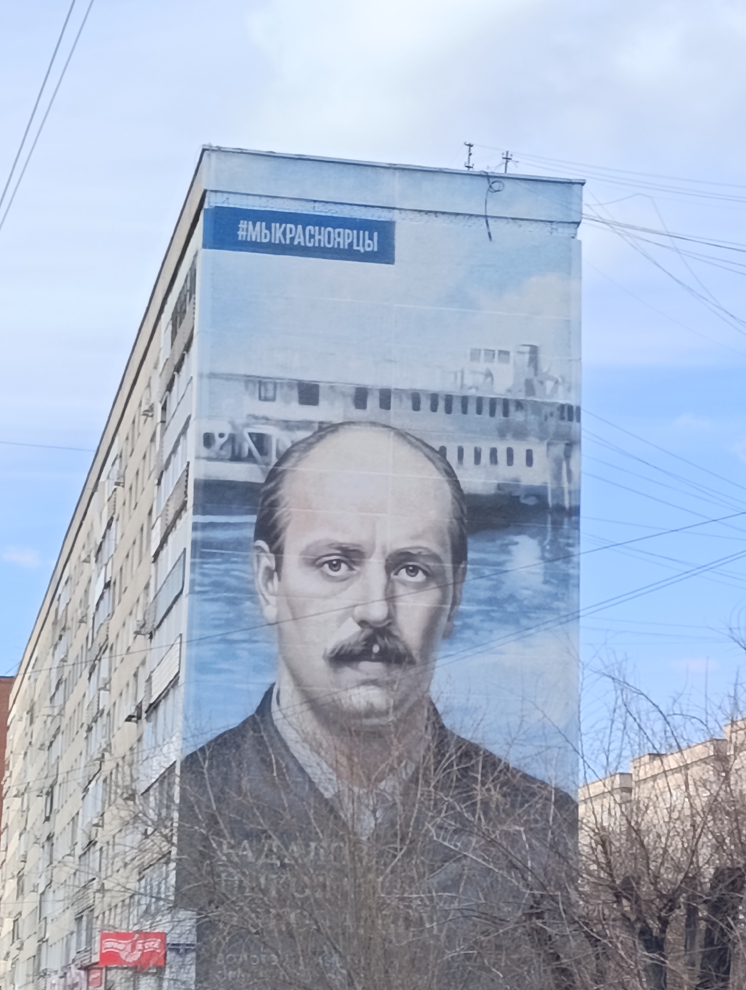 A large-scale mural adorns the entire side of a multi-story building, likely in Krasnoyarsk, Russia, as indicated by the hashtag. The mural features a prominent portrait of a man with a mustache and receding hairline, depicted from the chest up, with a neutral and focused expression. Behind him, a large, light-colored ship, possibly a steamboat or riverboat, is visible on calm blue water.
Above the portrait, a blue banner with white text reads "#МЫКРАСНОЯРЦЫ" (which translates to "#WEAREKRASNOYARSKERS" or "#WEAREKRASNOYARSKPEOPLE"). Faintly visible text below the man's portrait, partially obscured by bare tree branches, includes the name "Н.А. ДОЛГИХ" (N.A. DOLGIKH) and possibly "ВОЛГА" (VOLGA), referring to a ship or river.
The building itself is a tall, light-colored structure with numerous windows, suggesting a residential or office block. To the left, other similar buildings can be seen. The scene is set during daytime under a clear, light blue sky with some wispy clouds. Numerous thin overhead utility wires crisscross the upper right portion of the image. In the foreground, the bare branches of deciduous trees partially obscure the lower part of the mural and the street level, indicating a winter or early spring season.