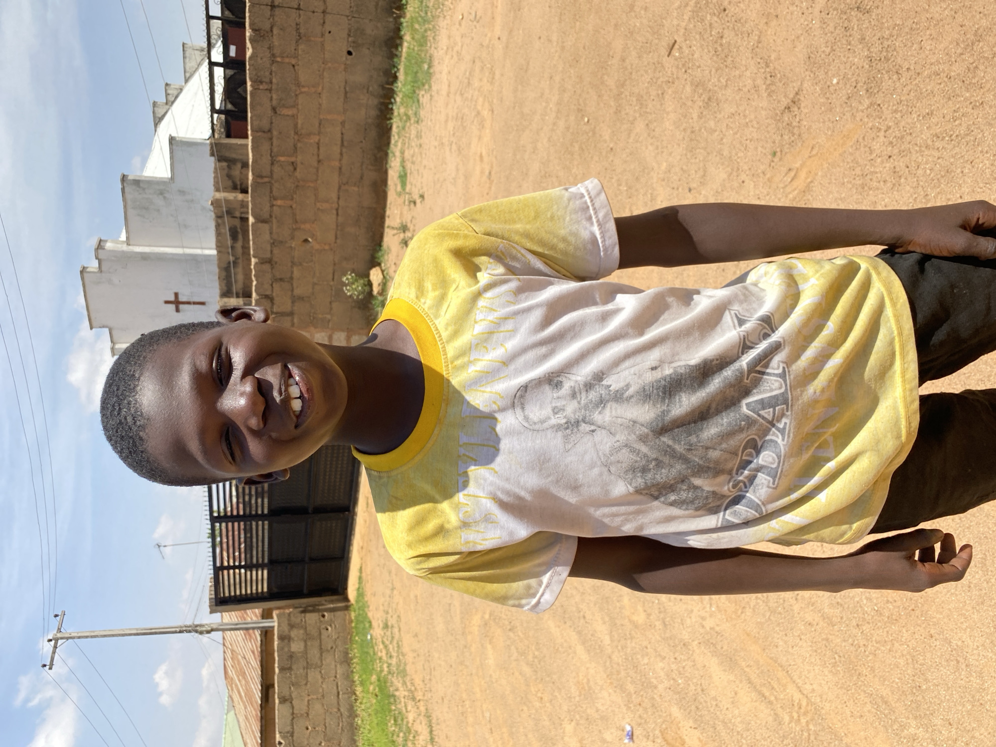 A young person smiles into the camera, wearing a yellow and white shirt and dark pants. The background shows a dirt ground and various buildings, including what appears to be a white church with a cross. An electrical pole is visible against a partly cloudy sky. The location is Bukuru, Nigeria.