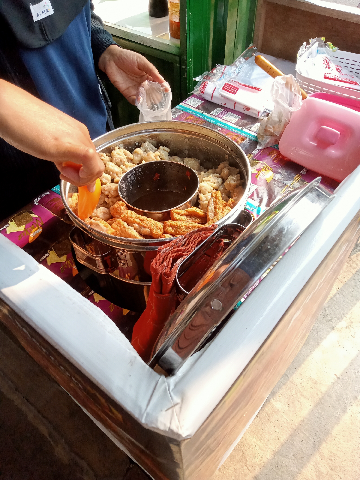In Simo, Indonesia, a vendor is preparing food at an outdoor stand. The vendor's hands are visible, one holding a plastic bag and the other using an orange utensil to select food items from a large metal pot. The pot contains a variety of fried snacks, including what appear to be meatballs and fish cakes, and a small bowl of dark sauce is placed in the center. The stand is covered with a colorful patterned cloth. To the right, there are packaged napkins and a pink plastic container. The scene is illuminated by natural sunlight, casting shadows on the ground.