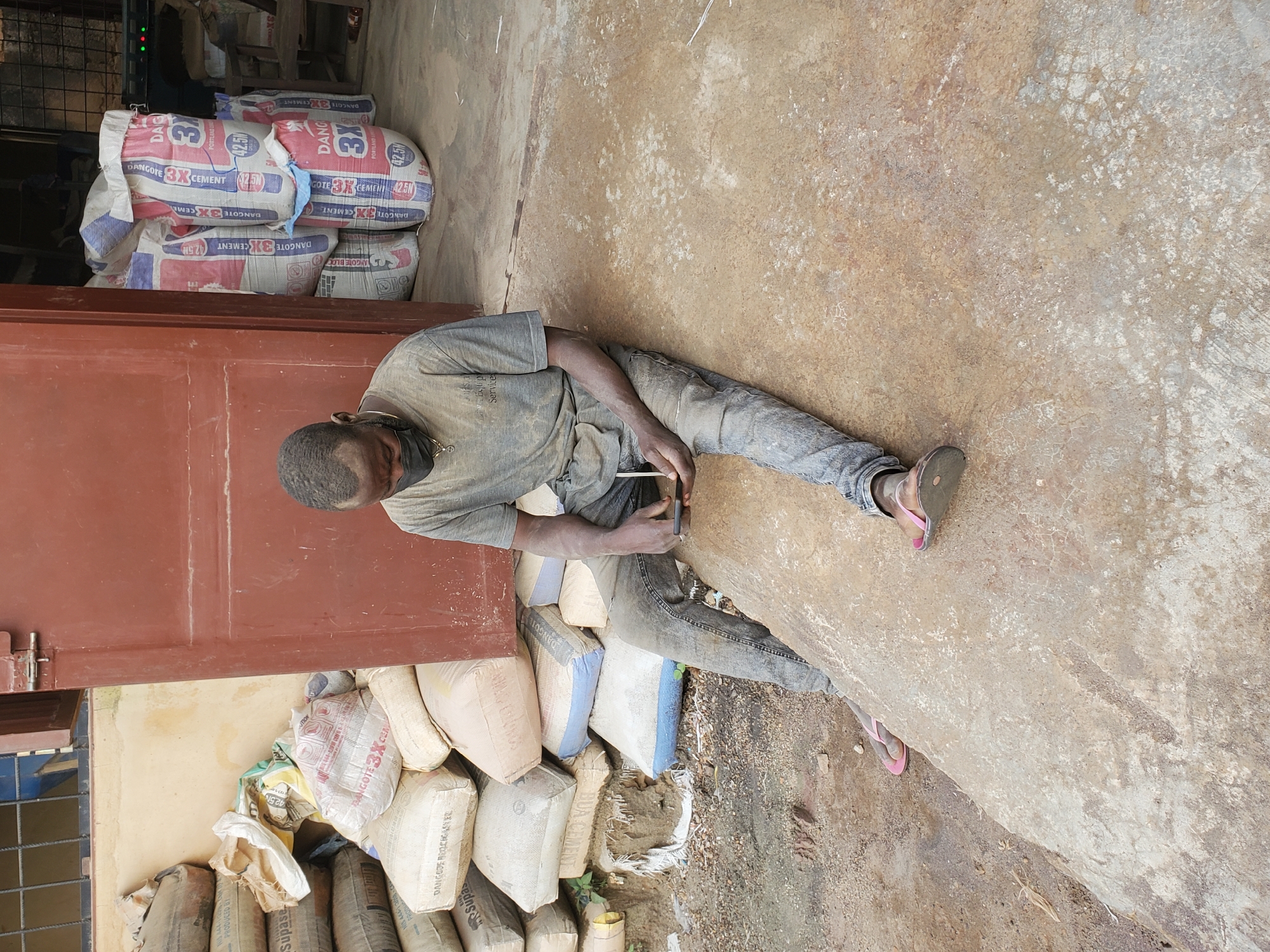 In Bukuru, Nigeria, a dusty, middle-aged Black man wearing a grey t-shirt, jeans, and a black face mask sits on a concrete slab outside a building with a red metal door. He is holding a smartphone in his hands and appears to be looking at it. Bags of cement are stacked behind him, some with "DANGOTE 3X CEMENT" printed on them. The ground is a mixture of dirt and concrete, with some scattered debris. The man's clothing and skin are covered in dust, suggesting he works in a dusty environment, likely related to construction or cement handling. The scene depicts a moment of pause and possibly rest during his workday.