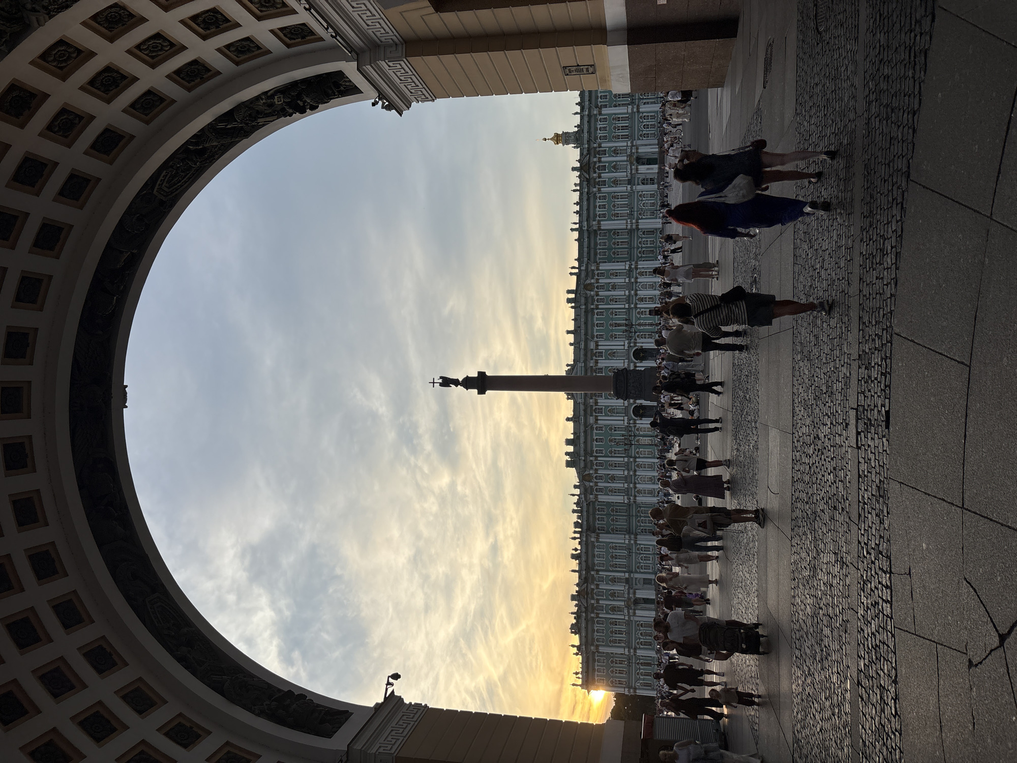 This is a ground-level, medium-shot, vertical-orientation photograph taken at Palace Square in Saint Petersburg, Russia. The view is framed by an archway, likely the General Staff Building, revealing a wide expanse of the square.
In the background, the Winter Palace, painted a distinctive pale green with white trim, stretches across the horizon under a hazy, sunset sky with scattered clouds. A tall, reddish-brown column topped with a statue of an angel stands in the middle ground, between the archway and the Winter Palace. The square itself is paved with cobblestones and large granite slabs.
A crowd of people are walking and standing in the square. Some are dressed in casual summer clothing. In the foreground, three women are walking away from the camera, two of them linked arm-in-arm. One woman is wearing a long blue dress, and the other a dark top and shorts. To their left, a man with a striped shirt and shorts walks away, carrying a bag over his shoulder. A stroller is visible to his left. The setting sun casts a warm glow, illuminating the scene and creating long shadows.