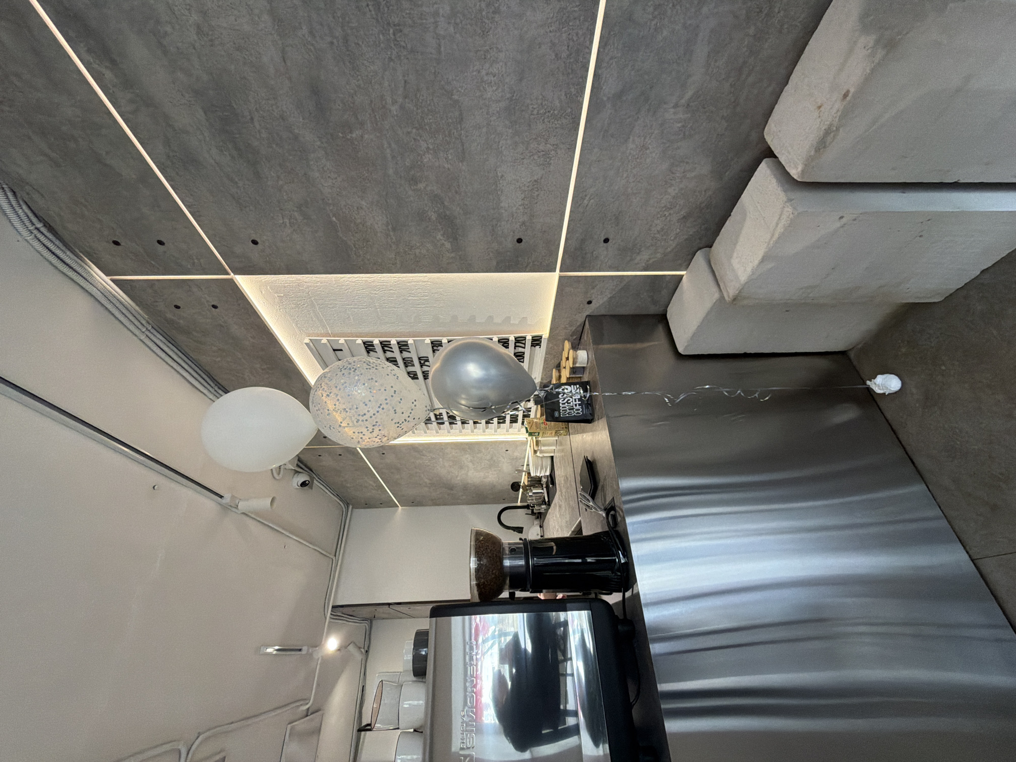 The image shows the interior of a business in Chaikovsky, Russia. The interior has a modern design with exposed concrete ceilings featuring linear lighting elements. Three balloons (white, silver and patterned) are visible. A stainless steel countertop dominates the right side of the image, reflecting ambient light. A Nuova Simonelli espresso machine is positioned on the counter. Shelving filled with cups is seen to the left.