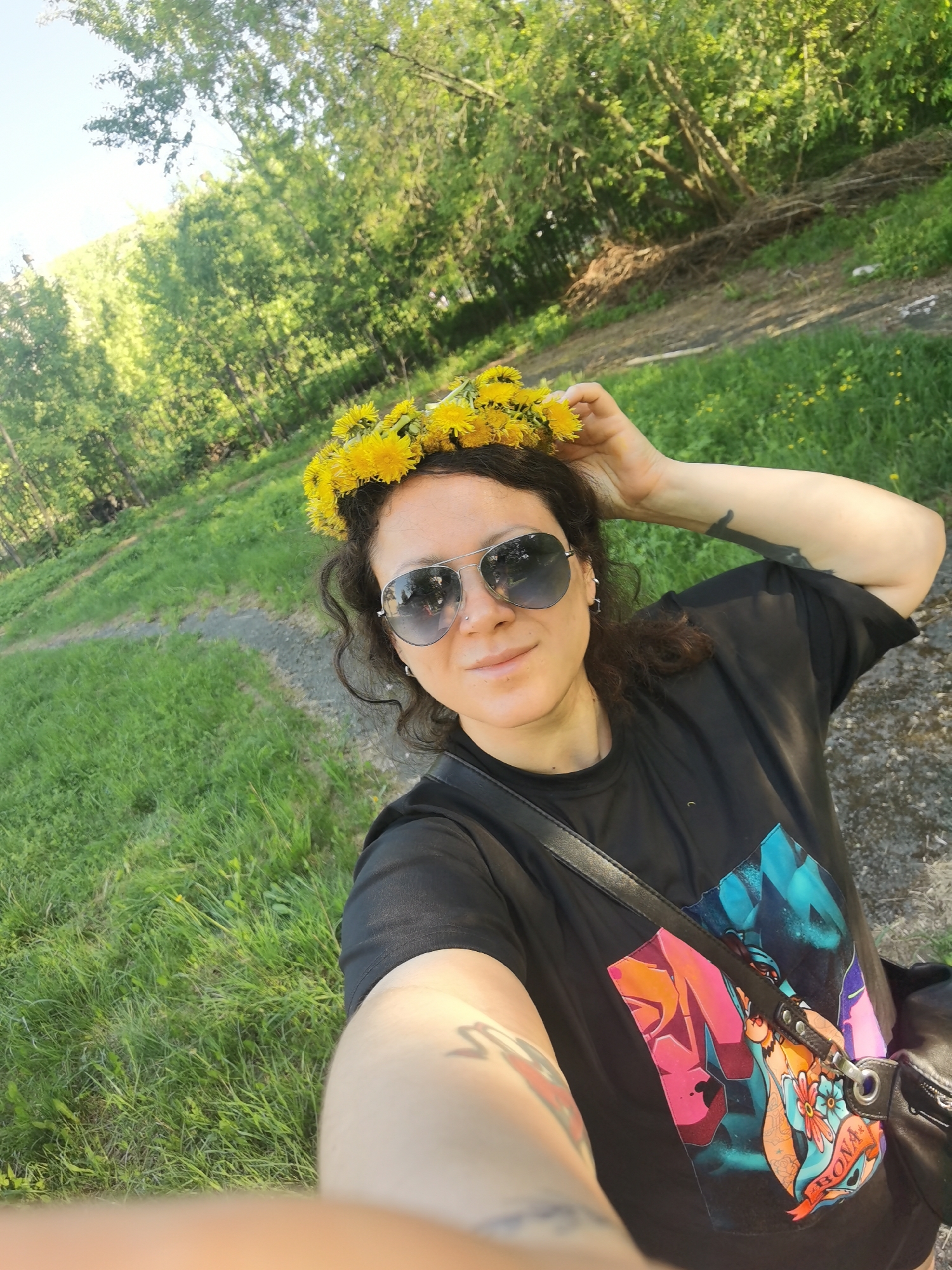 A person wearing a dandelion crown and sunglasses stands outdoors, taking a selfie on a sunny day in what appears to be a park or green space. The person is wearing a black t-shirt with a colorful graphic design and a black shoulder bag. The individual's hair is dark and curly. They are holding the dandelion crown with their right hand. Tattoos are visible on both arms. The background consists of grass, a gravel path, and trees. It is possible that the location is in Perm, Russia, although there are no visible landmarks to confirm it directly.