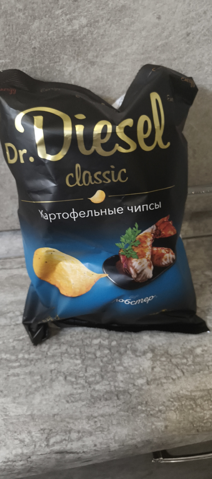 The image shows a bag of "Dr. Diesel Classic" potato chips. The bag is primarily black with gold lettering for the brand name and "classic" in a smaller font. Below that, in white lettering, it says "Картофельные чипсы," which translates to "Potato chips." A graphic of a garlic clove is positioned between the words "classic" and "Картофельные чипсы." The bottom portion of the bag features a blue gradient with an image of a potato chip and a small plate of food, possibly shrimp or prawns with herbs. The text "лобстер" (lobster) is visible on the blue part of the bag. The bag is placed on a textured, grey surface that appears to be a countertop or floor. The overall setting is an indoor environment, likely a home or store, given the plain background. The bag itself appears slightly crumpled, suggesting it has been opened or handled. There are no people visible in the image. The location in Aktanyshbash, Russia is not explicitly depicted or implied by the visual elements present.