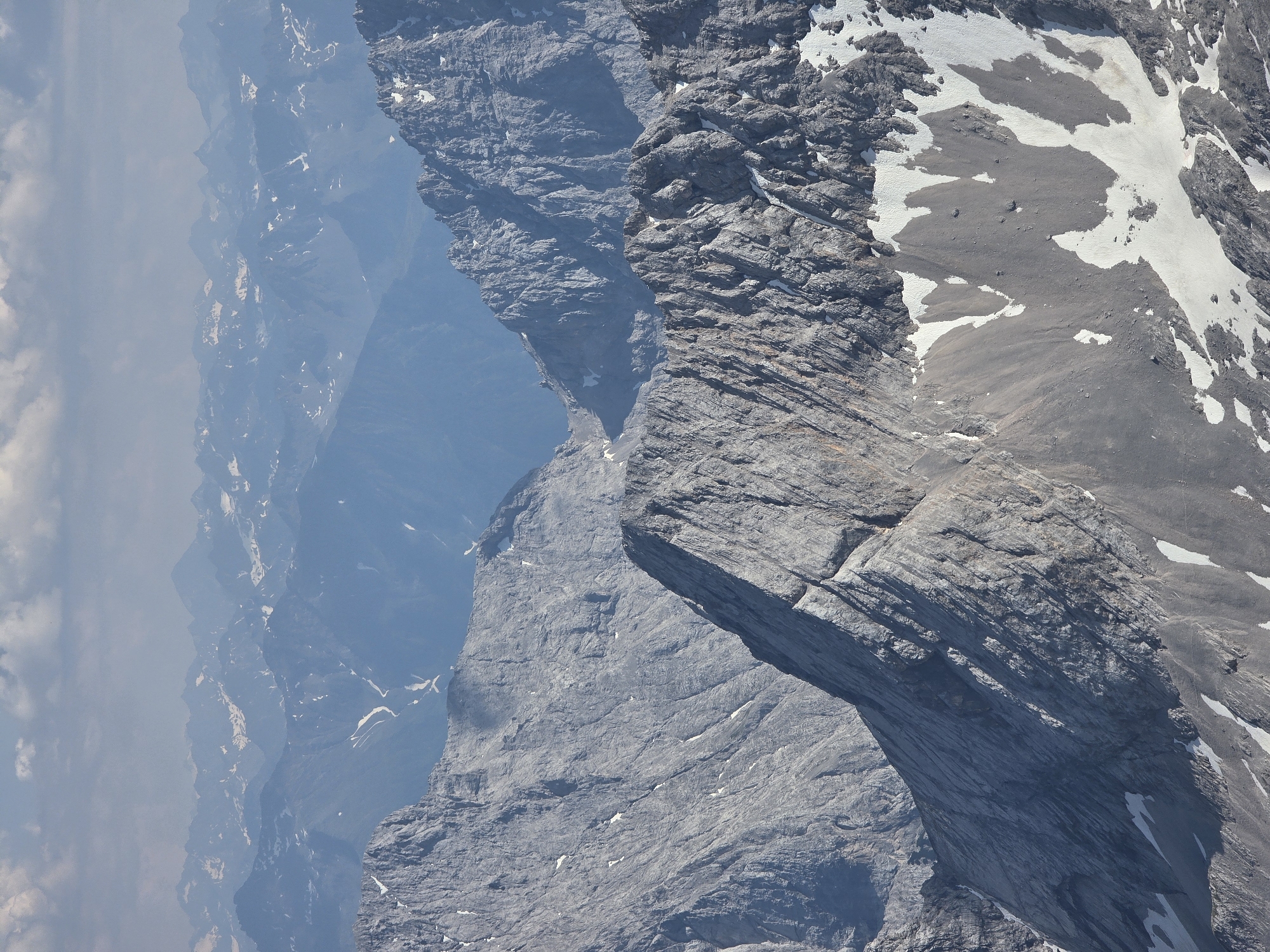 This is an aerial photograph of a vast mountain range, likely in Garmisch-Partenkirchen, Germany. The scene is dominated by rugged, gray, rocky peaks and slopes, with patches of snow scattered across the higher elevations, suggesting a cold, alpine environment. The lighting indicates it is daytime, with the sun casting shadows that highlight the dramatic contours of the terrain.
The view is from a high vantage point, looking down and across multiple layers of mountains that recede into a hazy blue distance. The foreground features a sharp, rocky ridge with distinct geological striations and overhangs. Further into the scene, softer, more undulating mountain forms are visible, partially obscured by atmospheric haze and clouds on the left side of the frame.
There are two small figures visible on a snowy slope in the middle ground, providing a sense of scale and indicating that people are present in this mountainous landscape, possibly hiking or mountaineering. The overall impression is one of grandeur, isolation, and the raw beauty of nature. The weather appears clear with some cloud cover, typical of a mountain environment. No text is visible in the image.