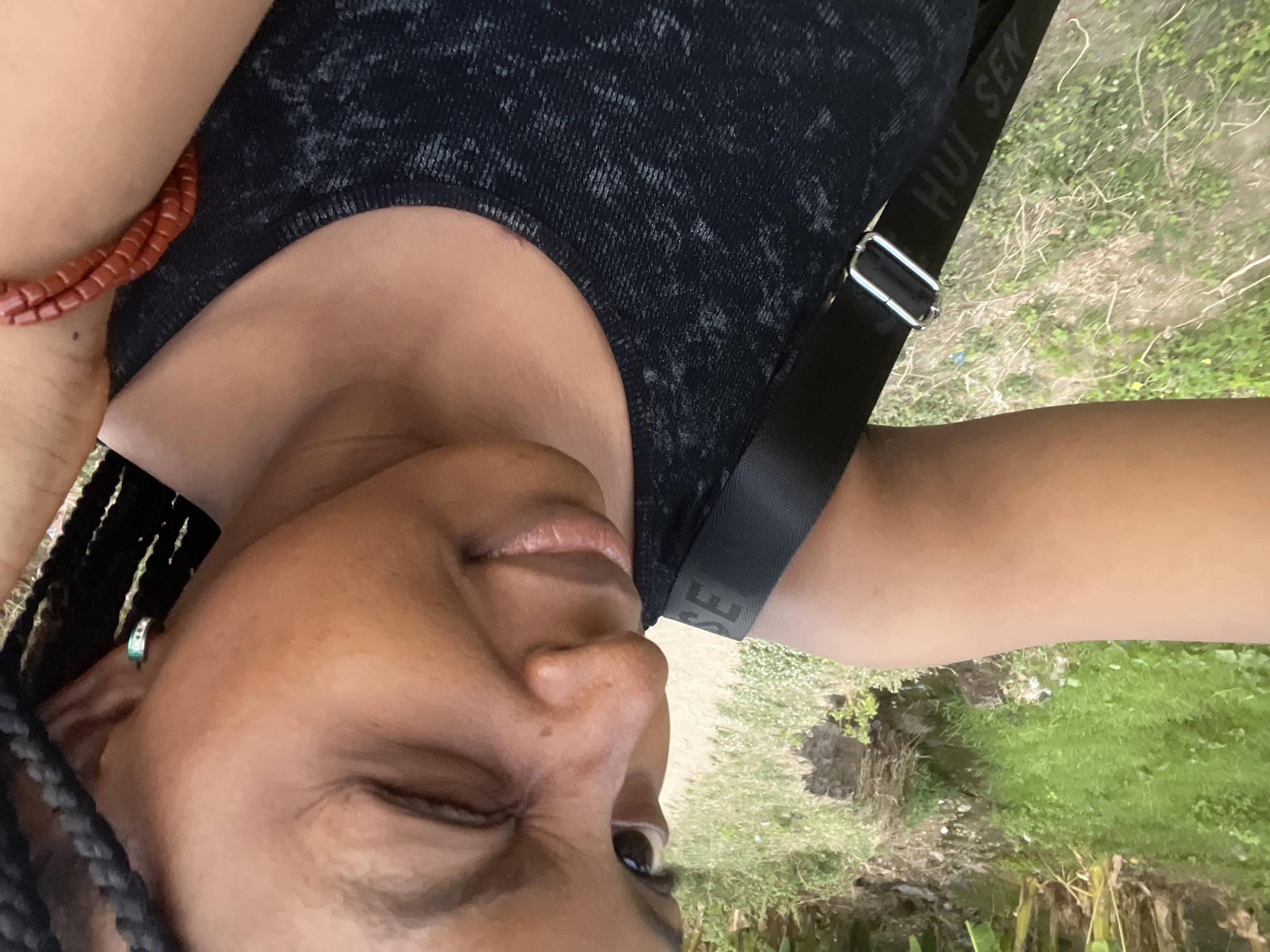 A close-up, tilted shot shows a woman outdoors. She is wearing a black top and a strap with the text "HUI SEN" across her chest. A beaded bracelet is visible on her wrist. The background shows a green, grassy area with dirt paths and vegetation. There is no specific evidence to place this scene in Lokoja, Nigeria.