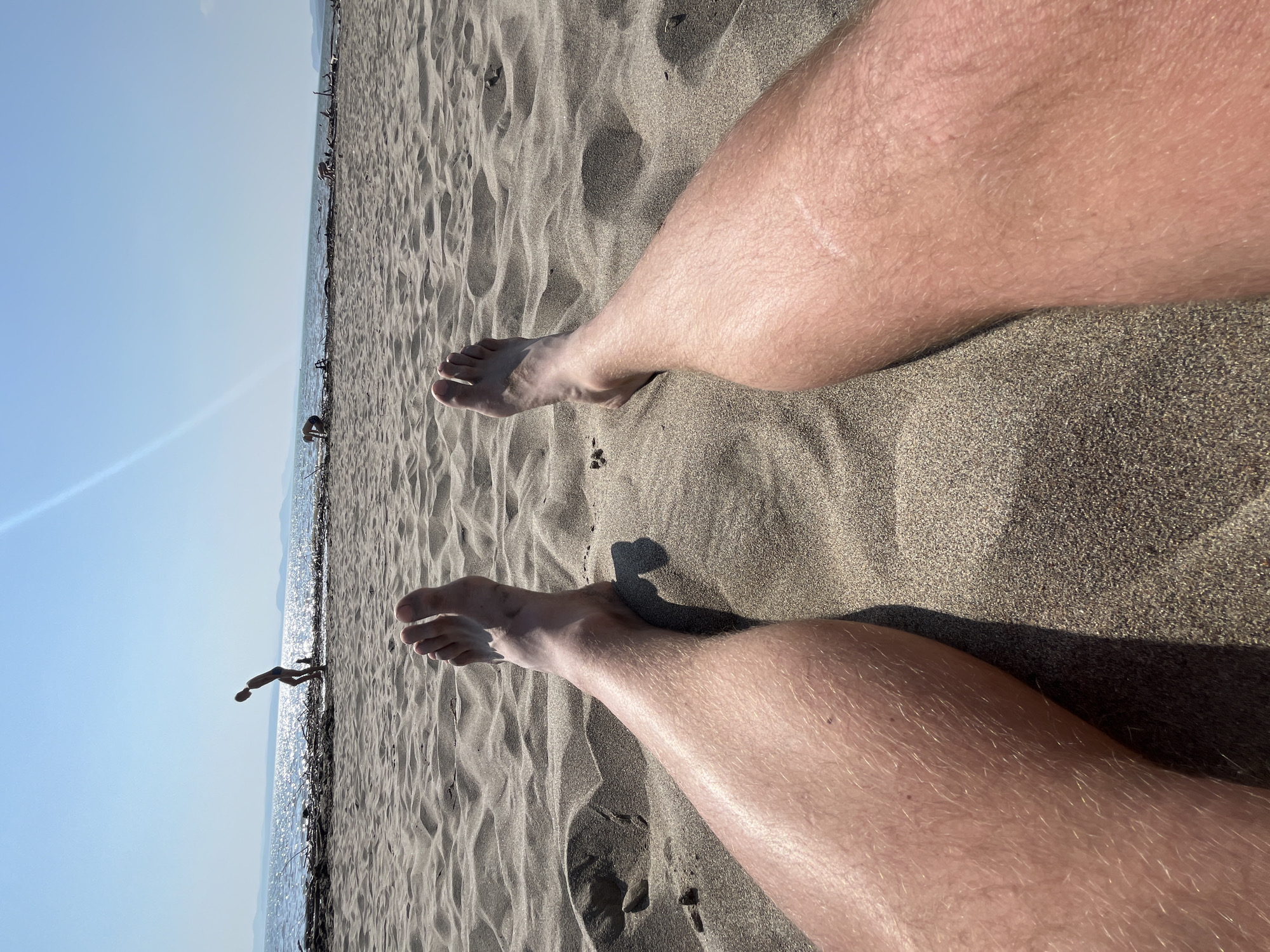 The image depicts a first-person perspective of a person relaxing on a sandy beach. The focus is on their legs and feet, which are partially buried in the sand. In the background, the sea is visible with gentle waves and a bright, sunlit sky. Two children are playing near the water's edge, one bending down and the other further in the distance. Several pieces of driftwood are scattered along the shoreline. The overall scene suggests a sunny day at the beach. The location is not identifiable as Krasnoturansk, Russia, or any other specific city based on the visual information provided.