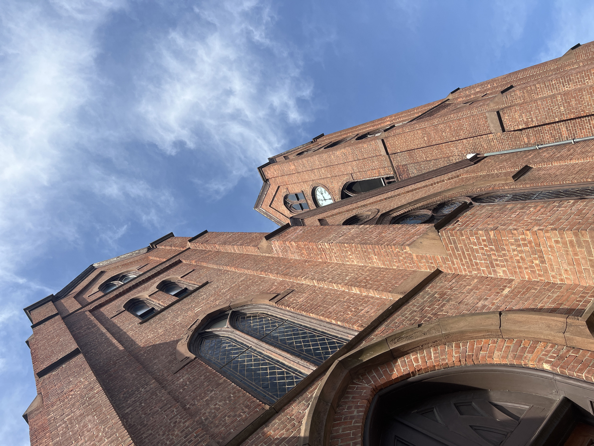 A low-angle shot captures a brick building with a sky backdrop in City of Newburgh, United States of America. The structure is constructed of orange-red bricks. A large arched window is situated above a door with a segmented panel design. Ascending upwards, a series of smaller, arched windows are also apparent. The sky above the building is blue with scattered light clouds. The composition is taken from a vantage point that emphasizes the building's height.
