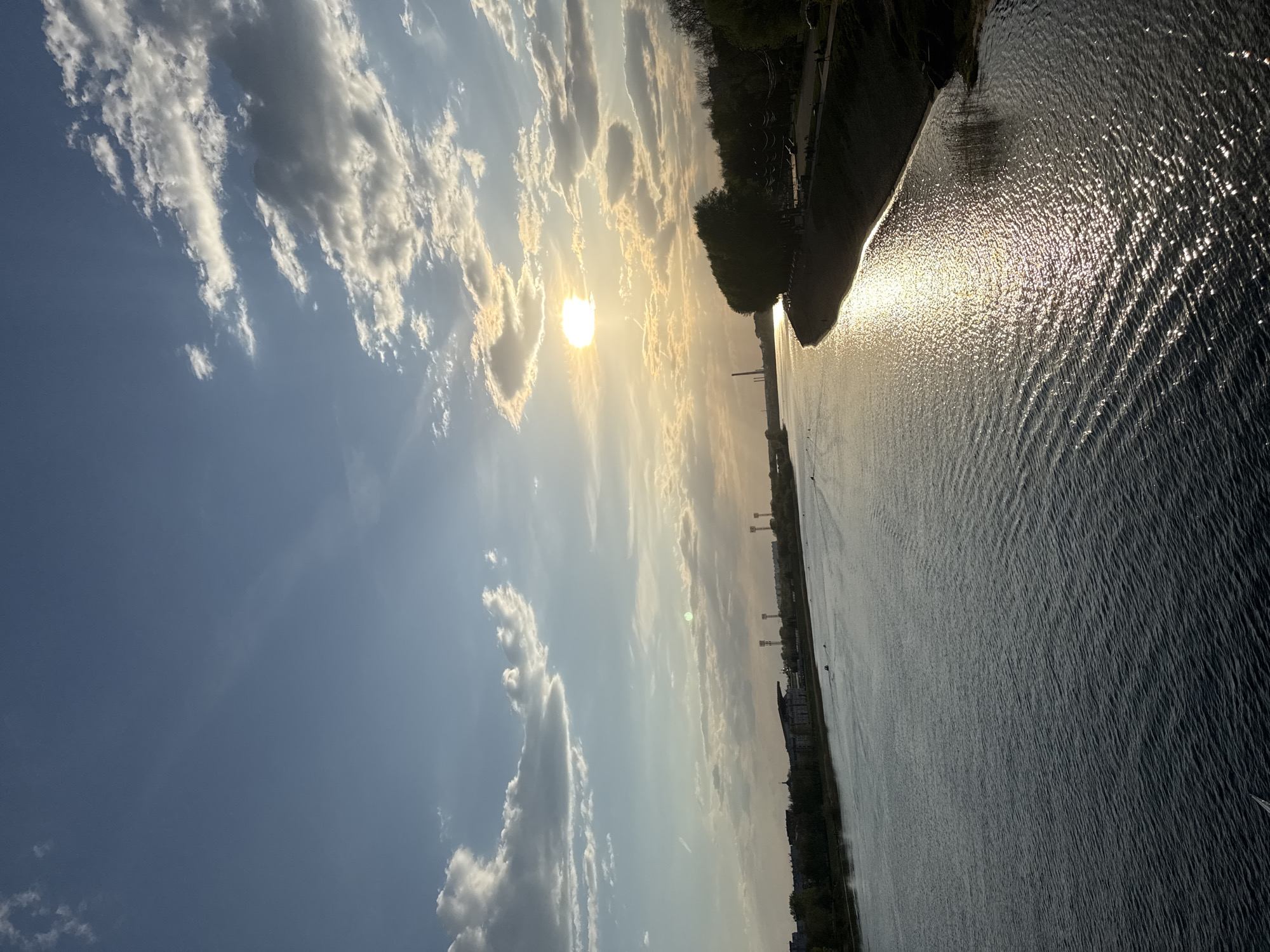 The image shows a view of the Volga River in Tver, Russia, at sunset. The sun is visible through the clouds in the sky, casting a bright reflection on the water. On the right side of the frame, the river's edge is visible, marked by a tree-lined bank. Several architectural structures, possibly buildings or towers, are seen further down the riverbank. The sky is partly cloudy with blue patches. The image is rotated to the side.