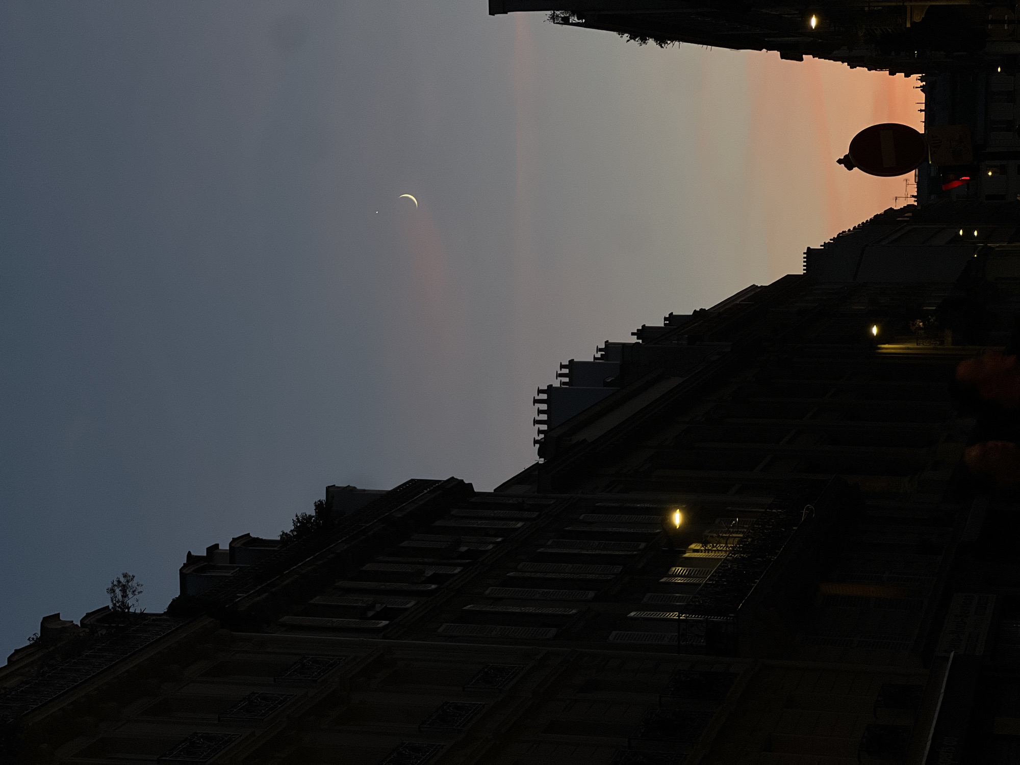 This is a vertical, low-angle shot of a narrow street in Paris, France, during twilight. On the left, a dark, multi-story building with ornate balconies and shuttered windows dominates the frame. To the right, another building of similar architecture is partially visible, also silhouetted against the sky. The street is dark, with only a few streetlights providing illumination. A red-and-white "no entry" sign is prominent on the right side of the street. The sky transitions from a deep blue at the top to a soft orange and pink hue near the horizon. A crescent moon and a bright star are visible in the upper right portion of the sky.