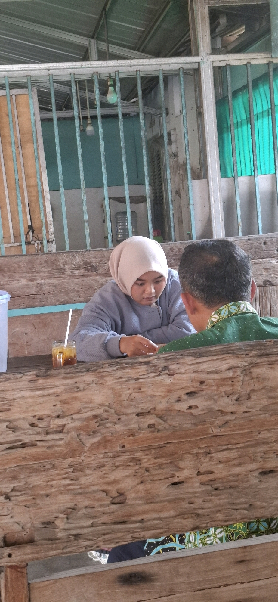 Two individuals, a woman wearing a light-colored hijab and a grey sweater, and a man wearing a green patterned shirt, are seated at a rustic wooden table. The woman is looking down, seemingly engaged with something on the table, with her hands positioned in front of her. A glass of iced tea with a straw is present on the table in front of her. The man is seated to her right, with his back partially to the camera, wearing a green shirt with a white and blue pattern. The background shows a weathered, turquoise-colored metal structure with vertical bars and some translucent green material. The overall setting appears to be an outdoor or semi-outdoor area, possibly a warung or eatery, with natural wood elements forming the table and seating. This scene is located in Pabelan, Indonesia.