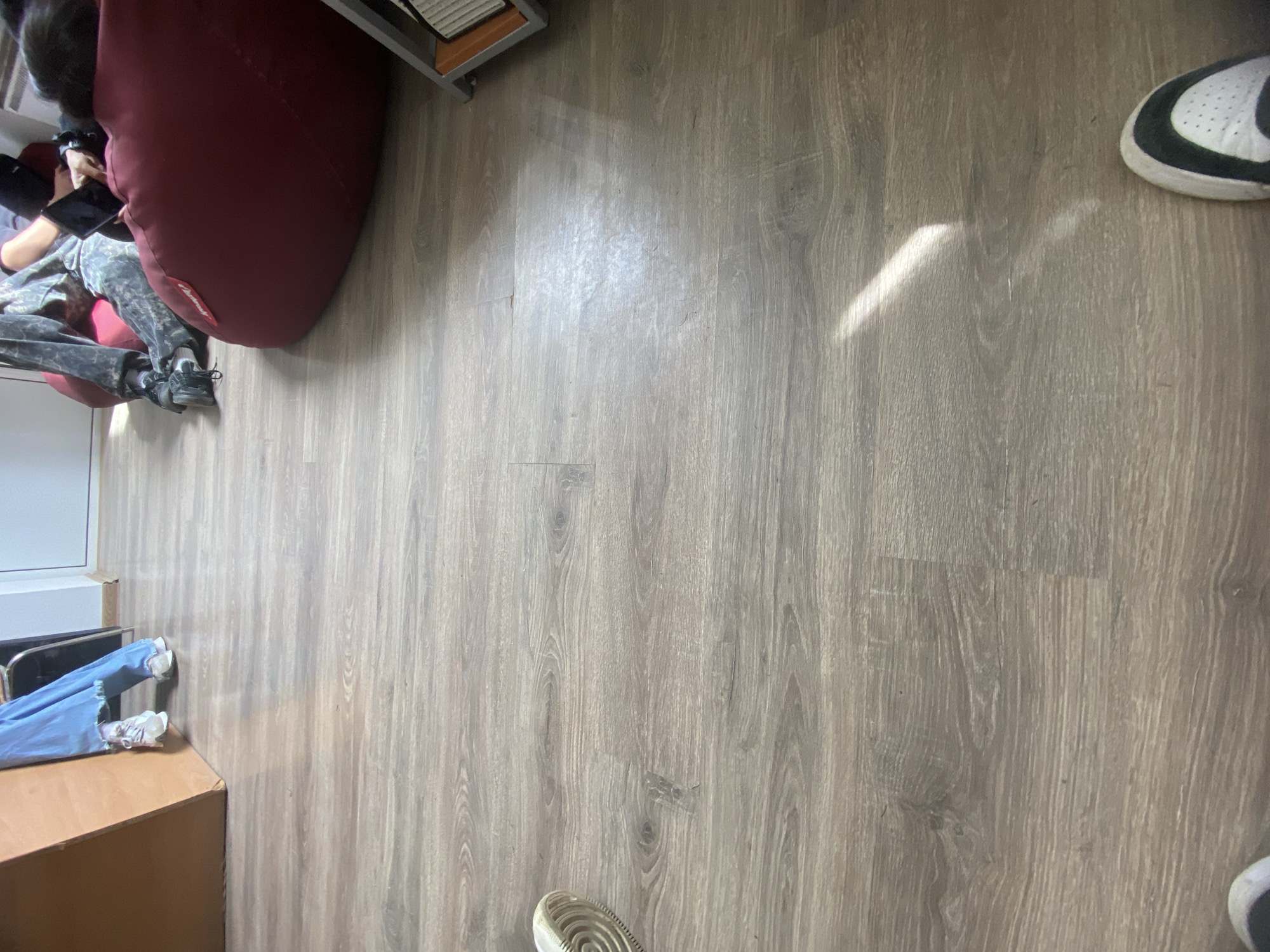 The image shows an indoor scene on a wooden floor. A person is sitting on a large red beanbag chair, holding a phone. Another person is lying on the floor near a desk, wearing jeans and white sneakers. A bookshelf is in the upper left corner. The location cannot be determined to be Troyan, Bulgaria.