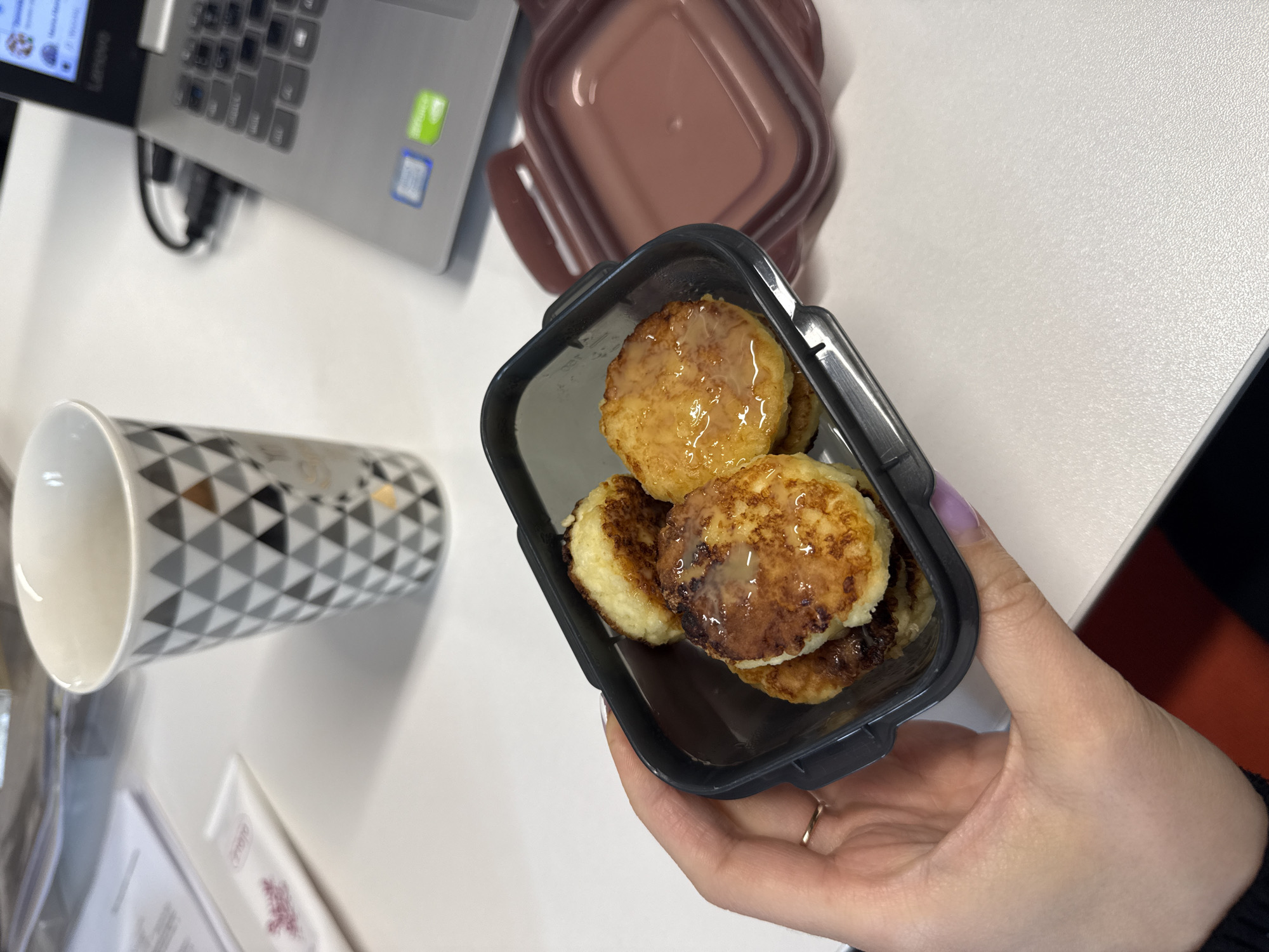 The image displays a close-up of a person's hand holding a dark grey food container filled with small, golden-brown, round fritters. The fritters appear to be syrniki or cheese pancakes, some of which are drizzled with a light-colored liquid, possibly condensed milk or syrup. The container has a transparent lid that is partially open.
In the background, a white desk surface is visible, supporting several objects. To the left, there is a white mug with a geometric pattern of black, grey, and gold triangles. Next to the mug, a white tube, possibly a cosmetic or sample product, is placed on some papers. Further to the right, a silver laptop is open, with its screen displaying content and a charging cable plugged in. A dark pink, square container lid rests on the desk to the right of the food container.
The overall scene suggests a casual setting, likely an office or a workspace, where a person is taking a break to eat. The lighting indicates an indoor environment. The image does not contain any specific landmarks or details that would allow for precise location identification within Łódź, Poland.
