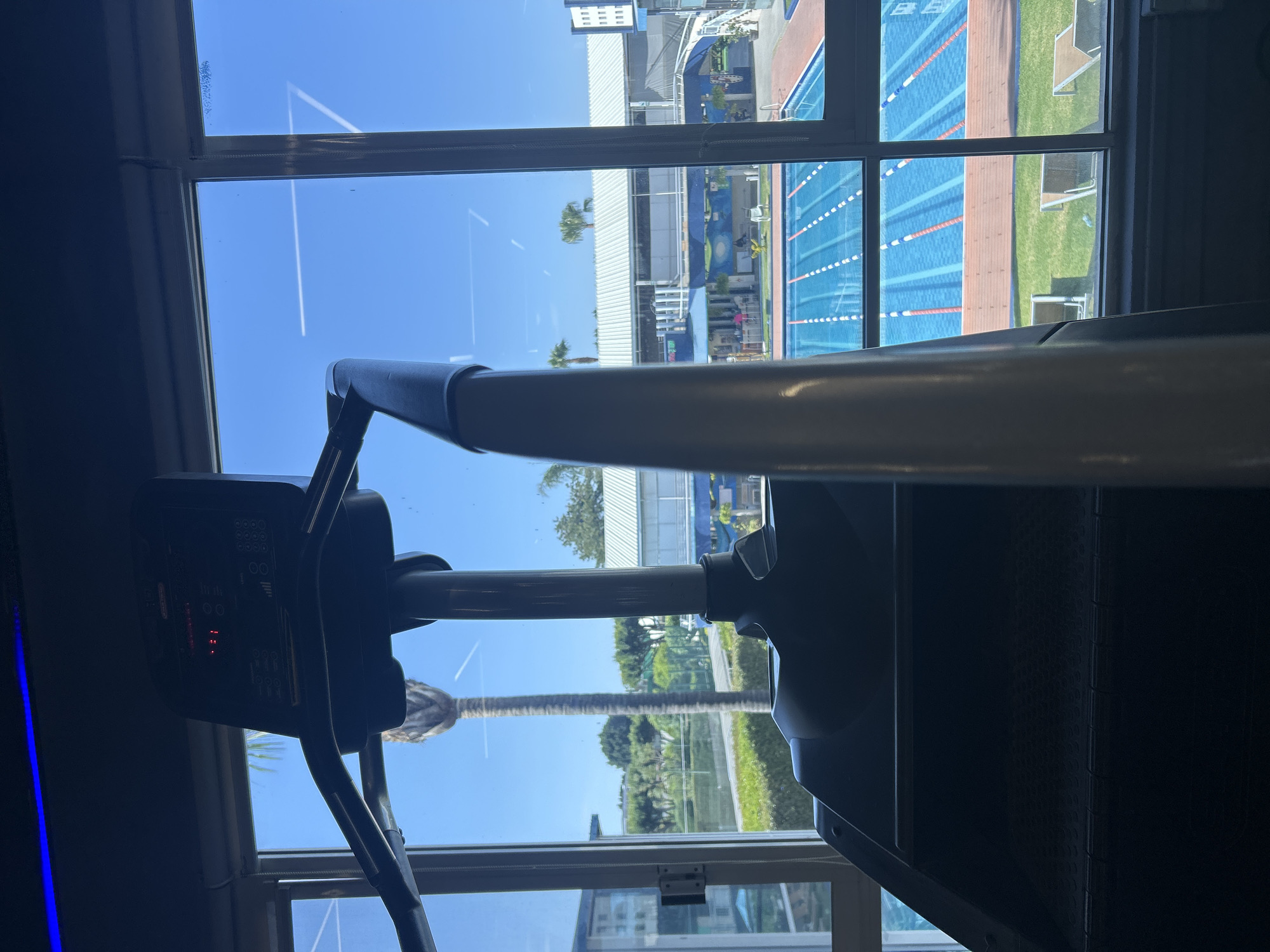 The image is taken from inside a building, likely a gym, in Puebla, Mexico. The focus is on a treadmill in the foreground, with the handles and control panel prominently visible. Through the window behind the treadmill, there is a view of an outdoor swimming pool area. The pool has multiple lanes marked with ropes, and there are lounge chairs and grass areas surrounding it. The sky is clear and blue, with visible contrails from an aircraft. The surrounding buildings appear to be part of a sports complex or recreational facility.