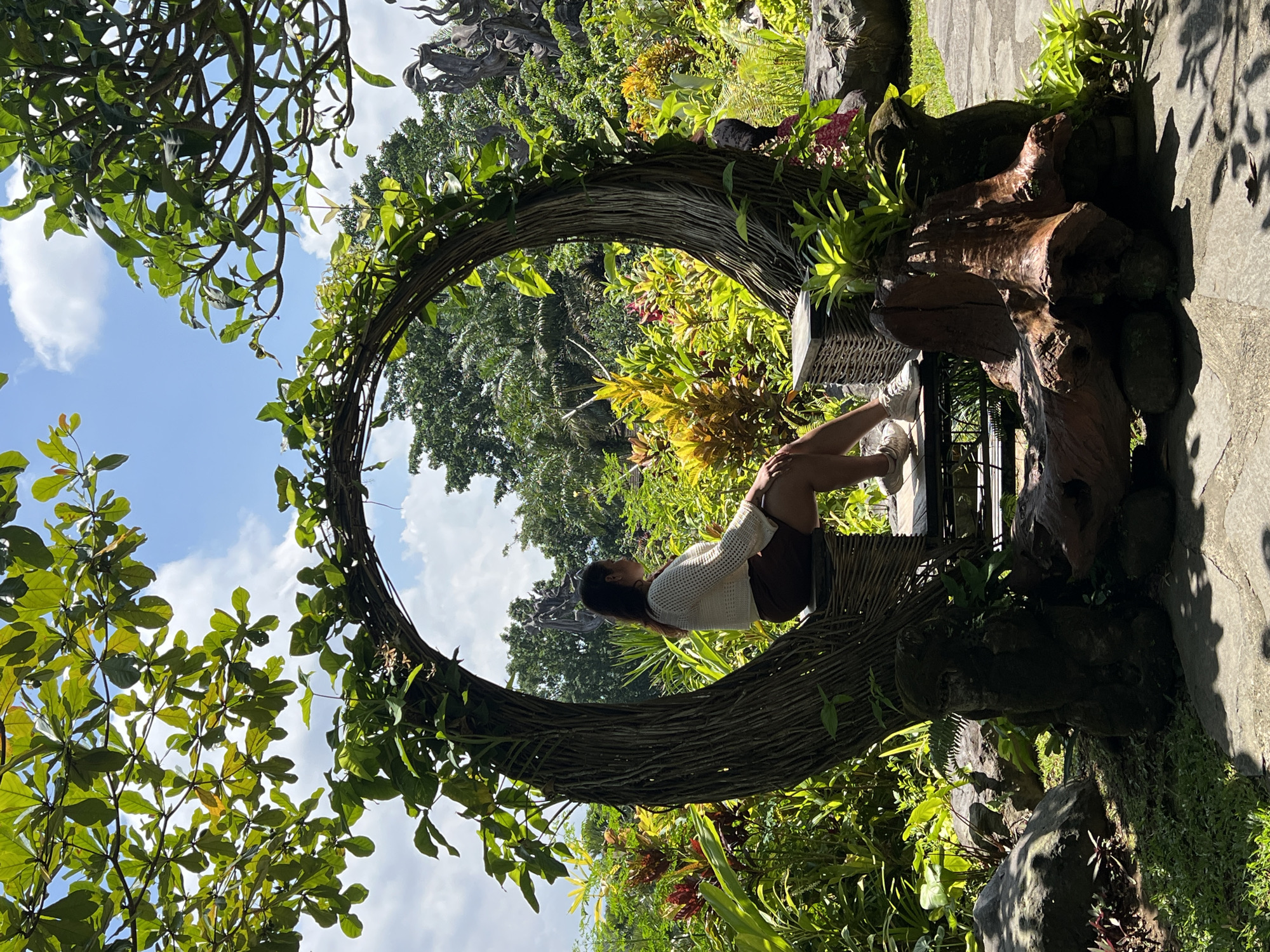 A woman is seated within a large, circular woven structure, integrated into a verdant garden setting. She has her legs bent towards her chest and is wearing a light-colored sweater and brown shorts. The woven structure appears to be supported by a wooden or faux-wooden base. The surrounding environment consists of lush greenery, including trees, plants, and flowers. The image captures a scene in Kedewatan, Indonesia.