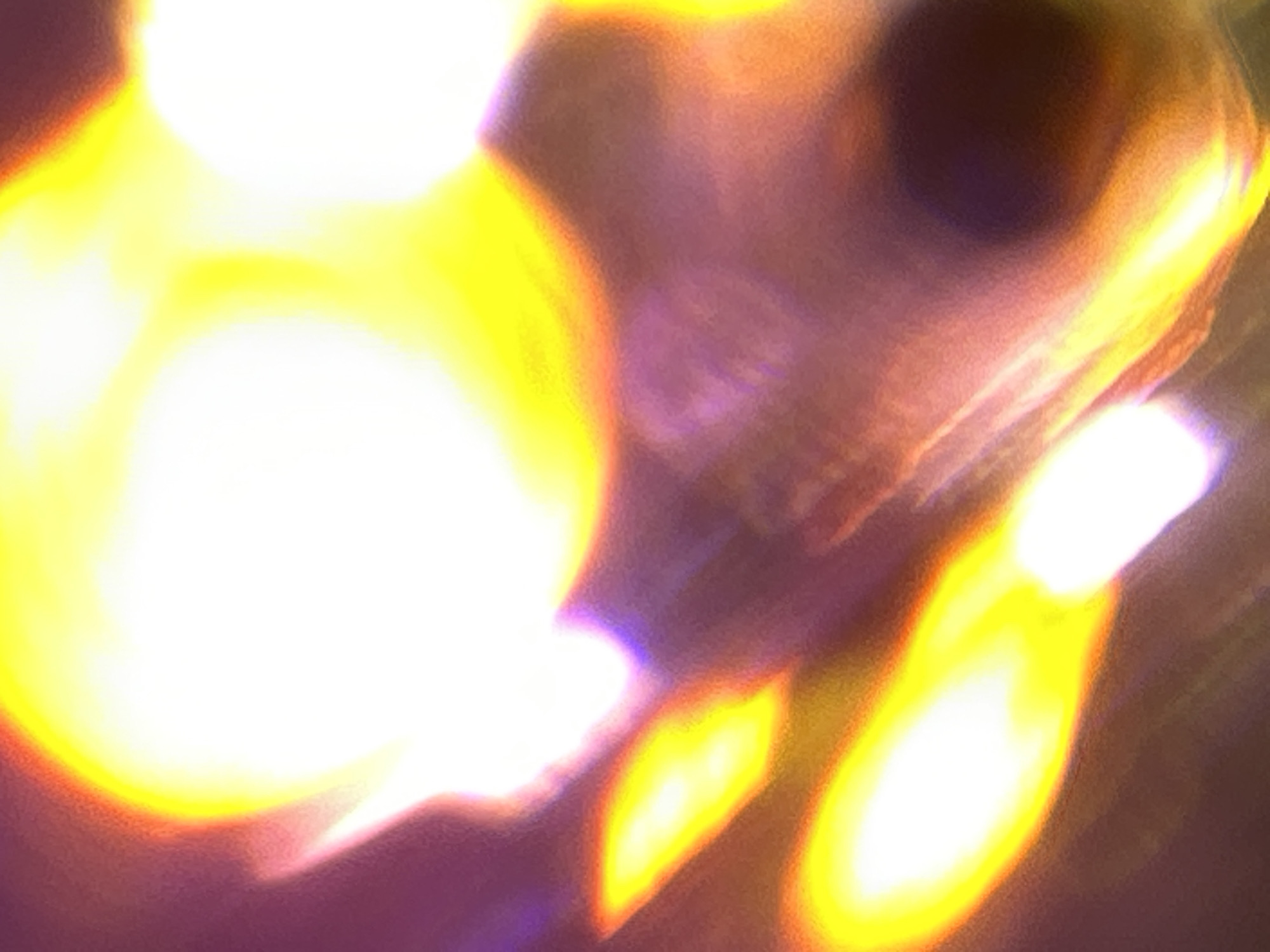 The image shows blurred yellow and white light sources against a dark background with hints of brown and purple. Some indistinct shapes may suggest objects, but they are not clearly discernible.