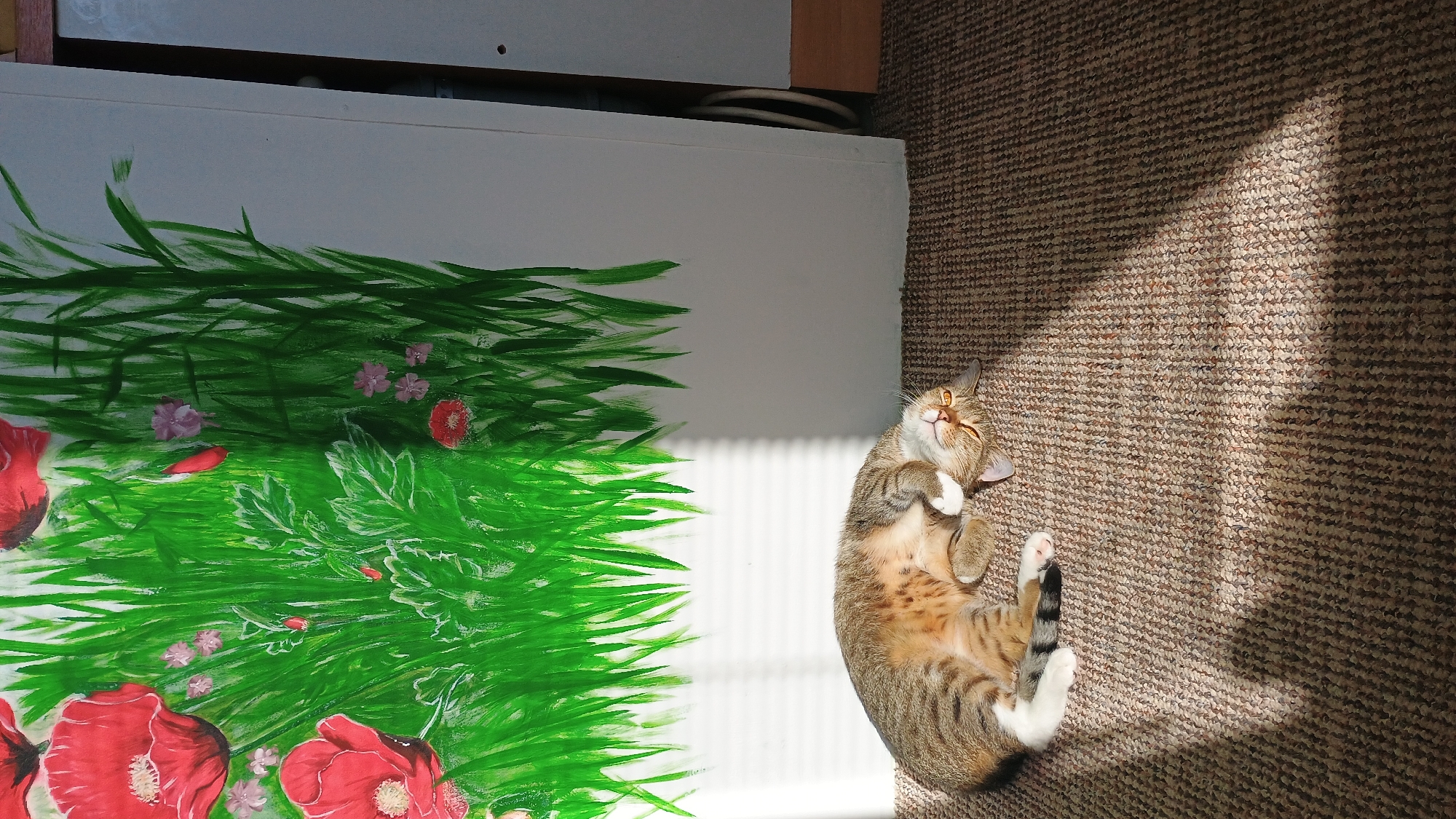 A tabby cat is sleeping on a carpeted floor in front of a white wall with a painting of green grass and red poppies. The sun is shining through a window with blinds, casting striped shadows on the wall and floor. The cat is curled up on its side, with its eyes closed and its paws tucked under its head. The painting appears to be a mural or a large canvas. The setting is indoors, possibly a living room or studio. There are no people visible in the image. The image is taken from a slightly elevated perspective, looking down at the cat. The overall lighting is warm, with bright sunlit areas and darker shadowed areas.