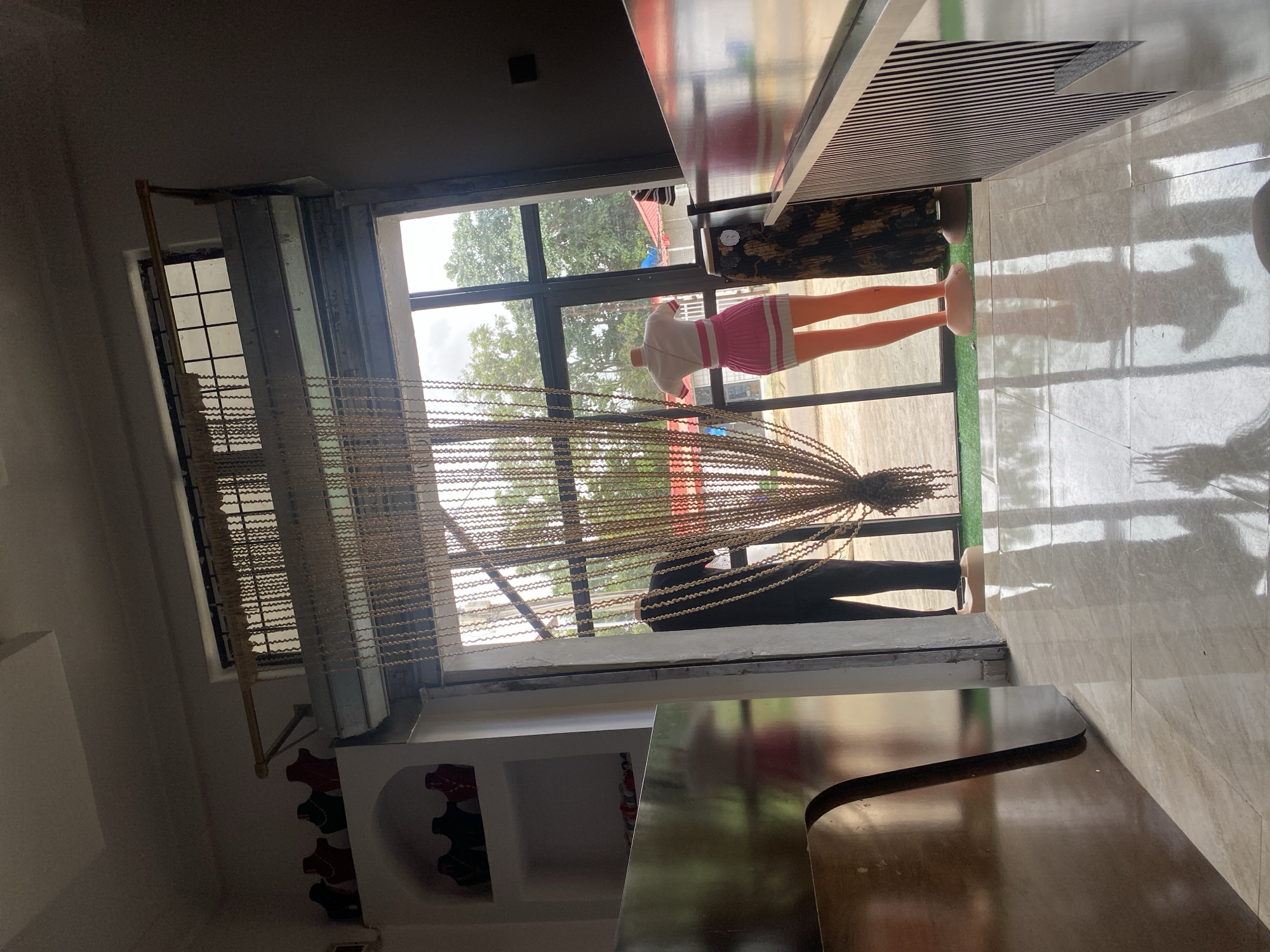 The image depicts the interior of a shop in Gura Top, Nigeria. The foreground shows a tiled floor with reflections from the overhead lighting. To the left, a large, curved wooden counter is visible, and beyond it, a shelf unit displays several black, red, and green jewelry busts, with more on a lower shelf. The middle ground features a doorway opening to the exterior, partially obscured by a long, beaded curtain. Through the curtain and doorway, two mannequins are visible. One mannequin, dressed in a black t-shirt and trousers, stands on the left. To its right, another mannequin wears a white and pink striped top and a matching pink pleated skirt with white trim. The mannequins stand on a green carpeted area outside the shop. Beyond the mannequins, trees and buildings with red roofs are visible through the window, suggesting an outdoor environment. The shop appears to be a clothing or accessory store.