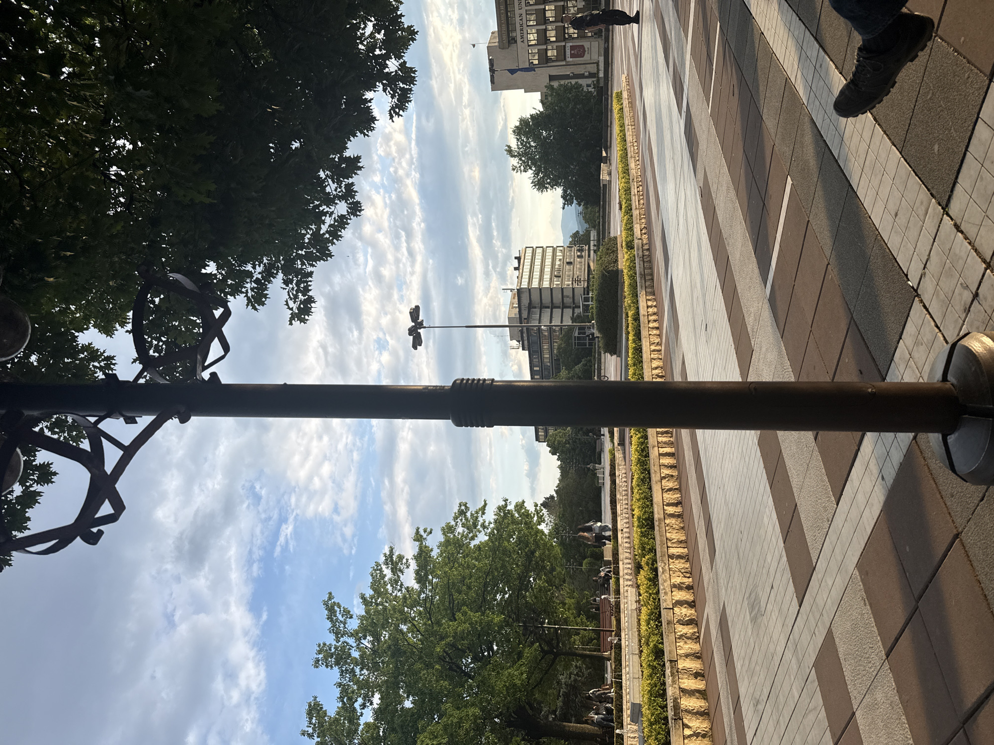 The image depicts an outdoor urban environment, likely a pedestrian square or pathway, viewed from a slightly low perspective. A prominent dark brown lamppost runs vertically through the left side of the frame, featuring an ornate, decorative structure at its top. The ground is paved with a distinct pattern of alternating light and dark grey/brown rectangular tiles, creating a striped and checkered design that recedes into the background on the right.
In the foreground on the far right, the legs and dark shoes of a person are visible, suggesting a close-up or first-person viewpoint. Lush green trees frame the scene, with dense foliage filling the upper left corner and another tree visible in the mid-ground on the right.
In the background, under a partly cloudy sky, several multi-story buildings are visible. One building prominently displays "AMERICAN UNIVER" on its facade, along with blue and white flags, indicating it is likely the American University in Bulgaria, aligning with the provided Blagoevgrad location context. Another modern building with numerous windows is situated further back. Low, well-maintained hedges with stone borders line the paved area.
The lighting suggests late afternoon or early morning, characterized by a soft, warm glow and visible shadows on the pavement, indicating a low sun angle. Several individuals can be seen walking casually in the background, engaged in everyday pedestrian activity. The overall mood is calm and unhurried.