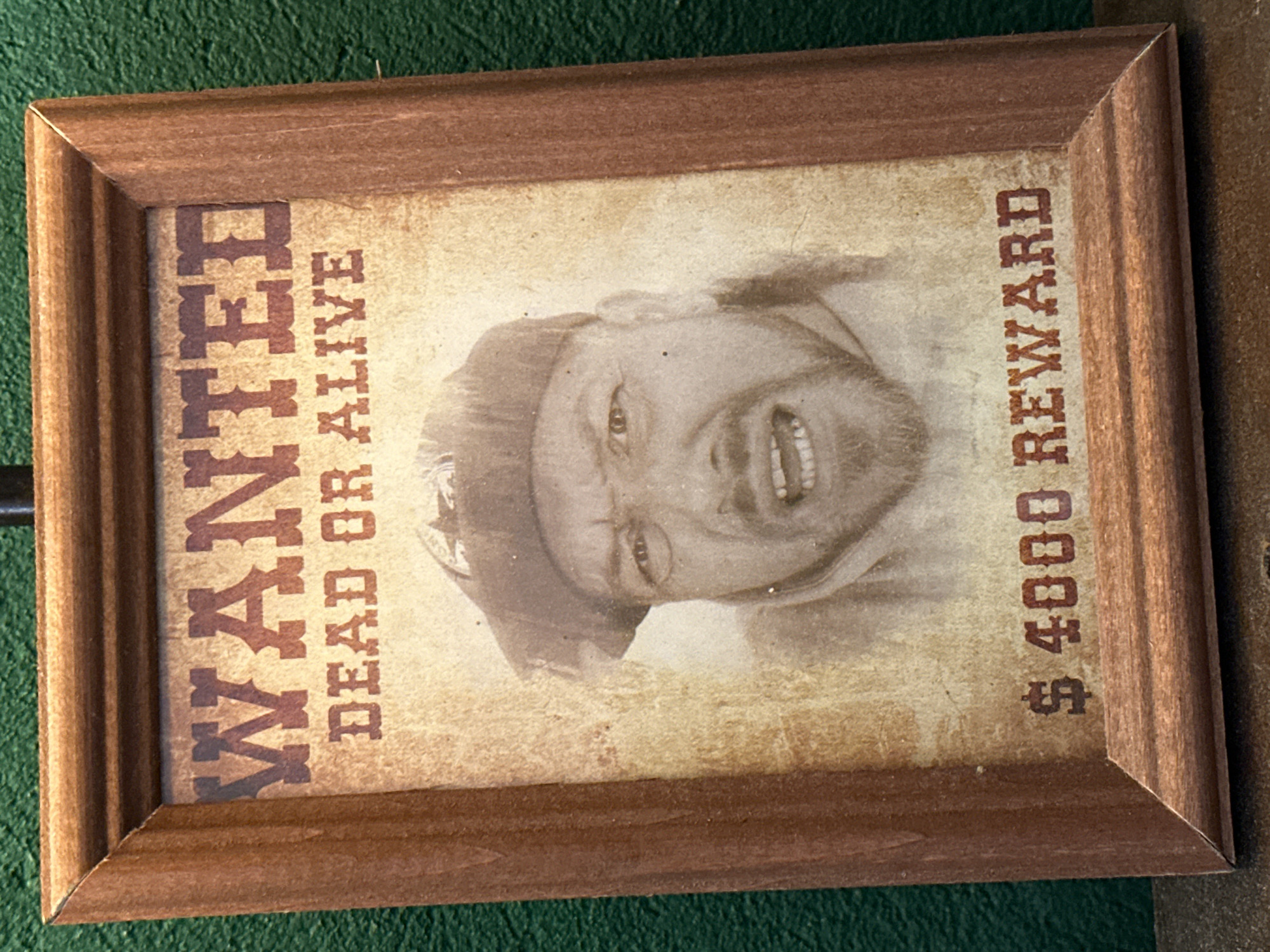The image shows a framed "Wanted" poster. The poster features a grayscale image of a man with a beard and a cap. Text on the poster reads "WANTED" on the left, "DEAD OR ALIVE" below "WANTED", and "$4000 REWARD" on the right. The poster has a distressed, aged appearance. The frame is made of wood. The poster is placed on a green surface. The location is not discernible from the image.