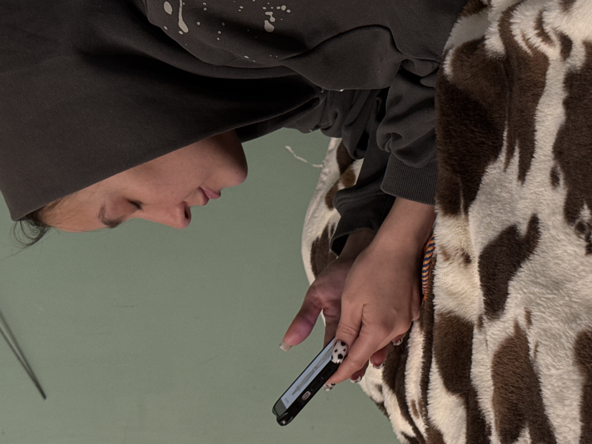 A woman wearing a dark grey hooded sweatshirt is looking down at a smartphone she is holding in her hands. She is sitting on a blanket with a brown and white cow print. The background is a light green surface. The location is not identifiable from this image.