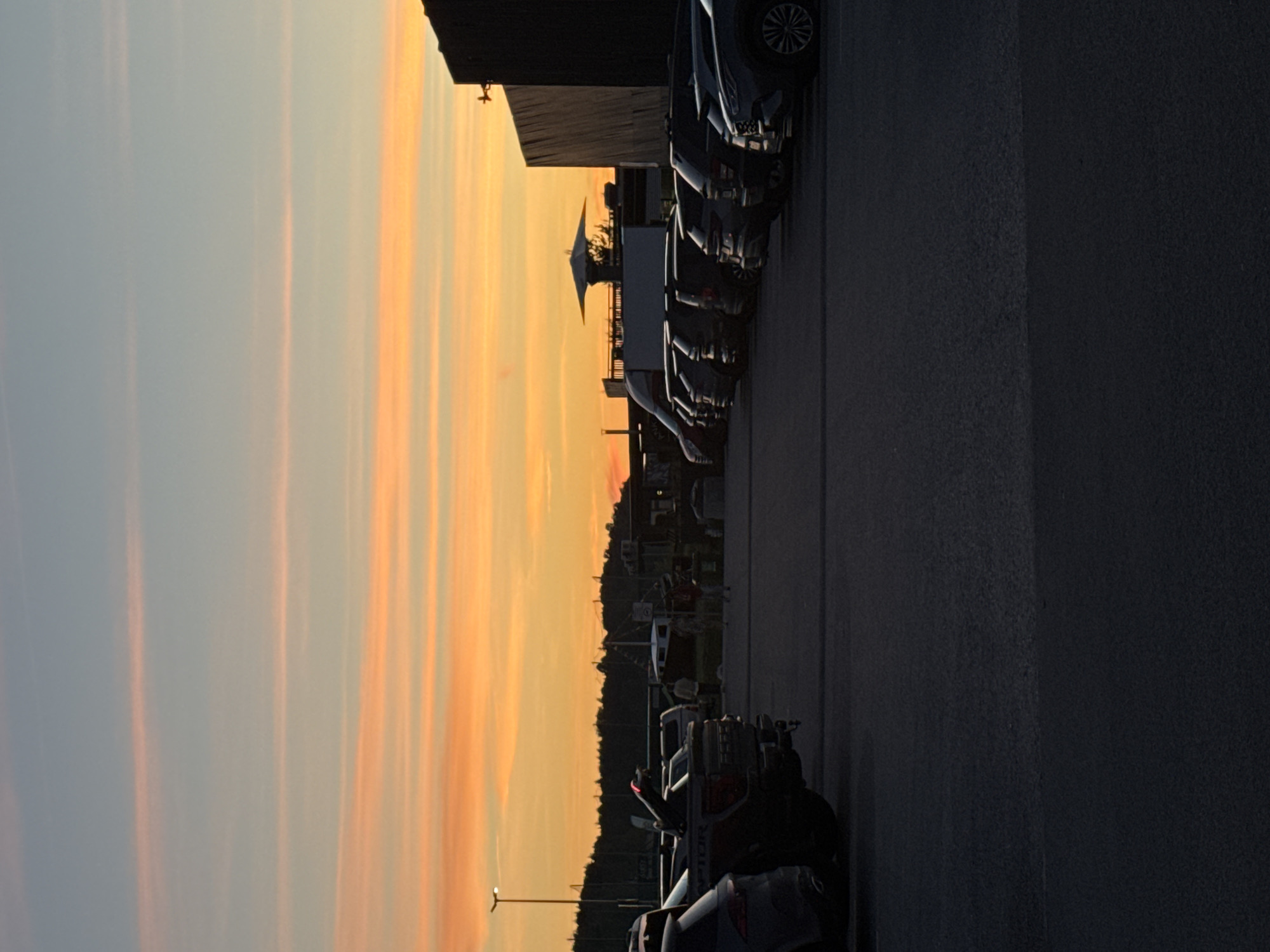 The image depicts a parking lot at sunset in Tallinn, Estonia. The sky is a vibrant gradient of orange and blue, with horizontal streaks of lighter orange clouds. In the foreground, several cars are parked. On the left, a dark-colored pickup truck is visible, with "RAPTOR" written on its side. To its right, other vehicles, including SUVs, are parked in rows.
In the background, silhouetted against the colorful sky, is a dark building, possibly a warehouse or industrial structure, with a flat roof and wooden-paneled walls. A distinctive, umbrella-shaped structure with spokes is visible on top of a lower building or structure to the left of the main building. Further in the distance, a line of trees forms a dark silhouette along the horizon. A street lamp is illuminated on the left side of the image. There are a few figures visible in the distance, appearing to walk in the parking lot. The overall scene suggests a commercial or industrial area on the outskirts of the city, captured during the twilight hours.
