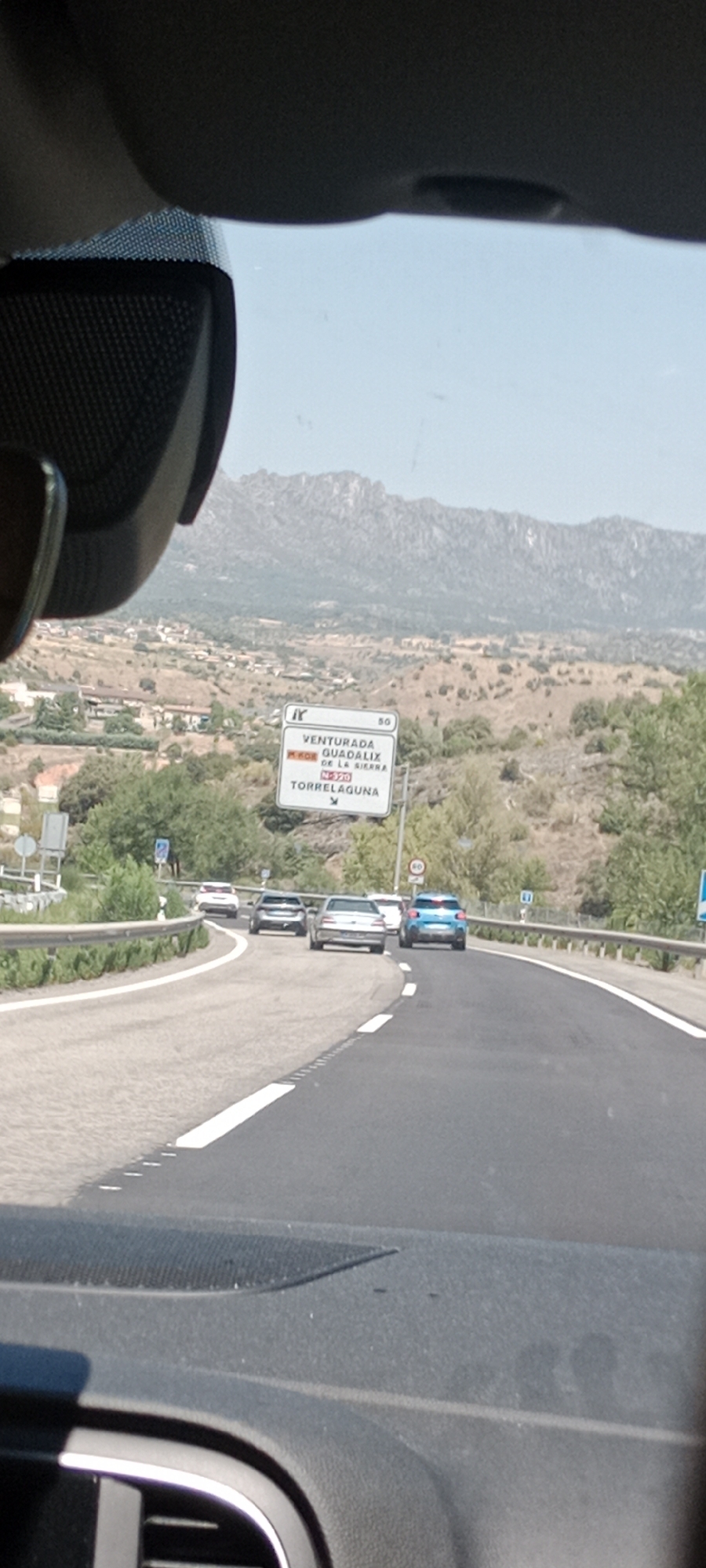 This is a first-person perspective shot from inside a vehicle driving on a road in Venturada, Spain. A sign is visible to the right of the road, indicating "VENTURADA GUADALIX DE LA SIERRA" and "TORRELAGUNA" with road numbers "M-608" and "N-320". Several cars are driving on the road ahead. The landscape is hilly and dry, with sparse vegetation and some buildings visible in the distance. The sky is clear and blue. The dashboard and windshield of the vehicle are partially visible in the foreground.