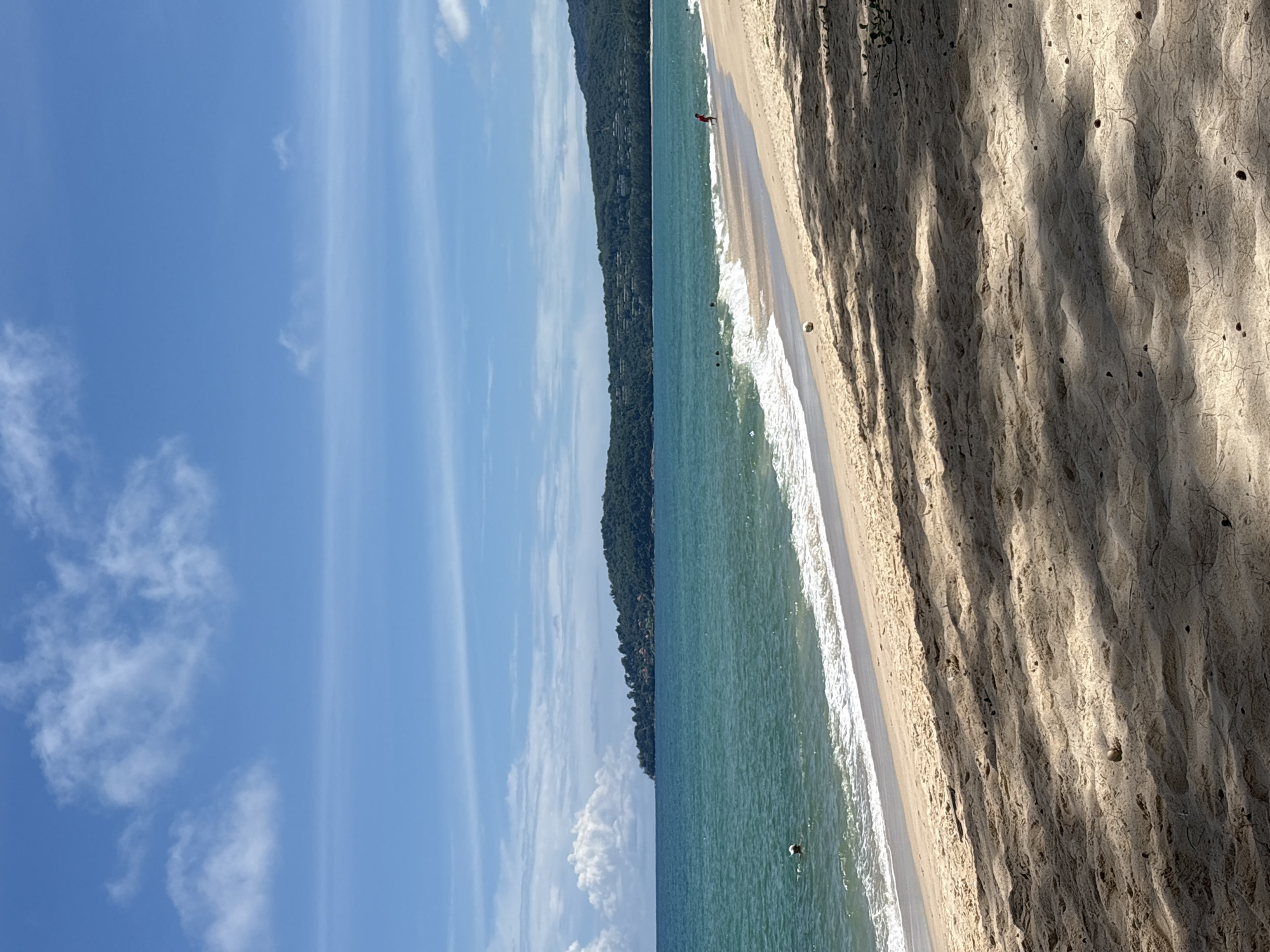 A wide, sandy beach in Choeng Thale, Thailand, stretches from the foreground into the distance. The sand is light brown, with shadows from palm trees dappling the surface, creating diagonal patterns. A gentle wave breaks on the shore, its white foam creating a distinct line between the water and the sand. The ocean is a clear turquoise, with small ripples and a few scattered swimmers visible. In the distance, a lush, green, tree-covered hillside rises from the sea, dotted with buildings. The sky is a vibrant blue, with scattered white clouds and a faint haze.