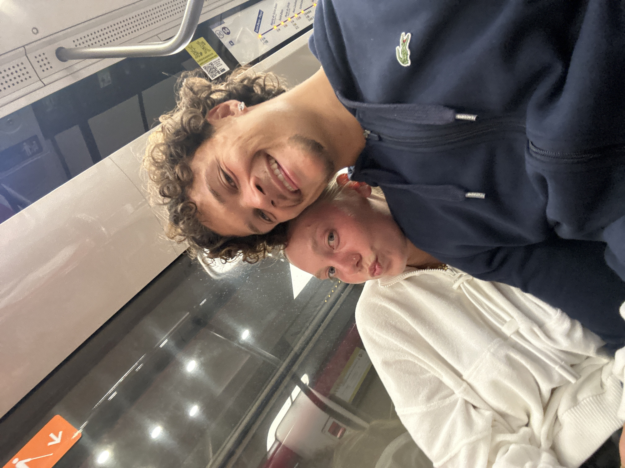 Two people are posing closely together inside a train or subway car, possibly in the city of Paris, France. A man with curly hair and wearing a dark blue Lacoste jacket is on the left with a broad smile. The other person, possibly a woman, is wearing a white jacket and has a pursed expression. The train car interior is visible in the background, with a window, handrail, and signage visible.