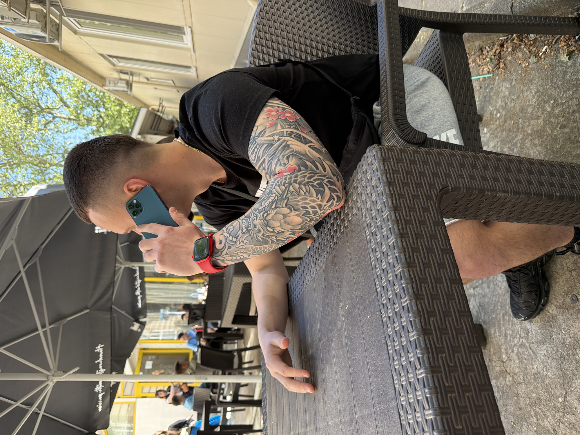 This media file captures a man seated at an outdoor cafe in Plovdiv, Bulgaria, talking on a blue smartphone. He is positioned sideways, leaning on a dark brown woven plastic table, with his left arm resting on it. His right arm, which is closer to the camera, is covered in a prominent and intricate full-sleeve tattoo featuring a large, stylized dragon or creature intertwined with red cherry blossom flowers.
The man has short, faded hair and is wearing a black t-shirt, a gold chain around his neck, grey sweatpants (with "NIKE" partially visible on the leg), and black sneakers. A red smartwatch is visible on his left wrist.
The setting is an outdoor area, likely a pedestrian zone or terrace. In the background, several large black outdoor umbrellas with unreadable white text on them are visible, along with other dark woven plastic tables and chairs. Other patrons can be seen blurred in the distance, suggesting a busy environment. Beyond the seating area, green trees with lush foliage are under a clear blue sky, and a building with windows and a distinctive yellow trim or awning is visible. Air conditioning units are mounted on the building wall. The ground is a light-colored concrete surface. The scene appears to take place on a bright, sunny day.