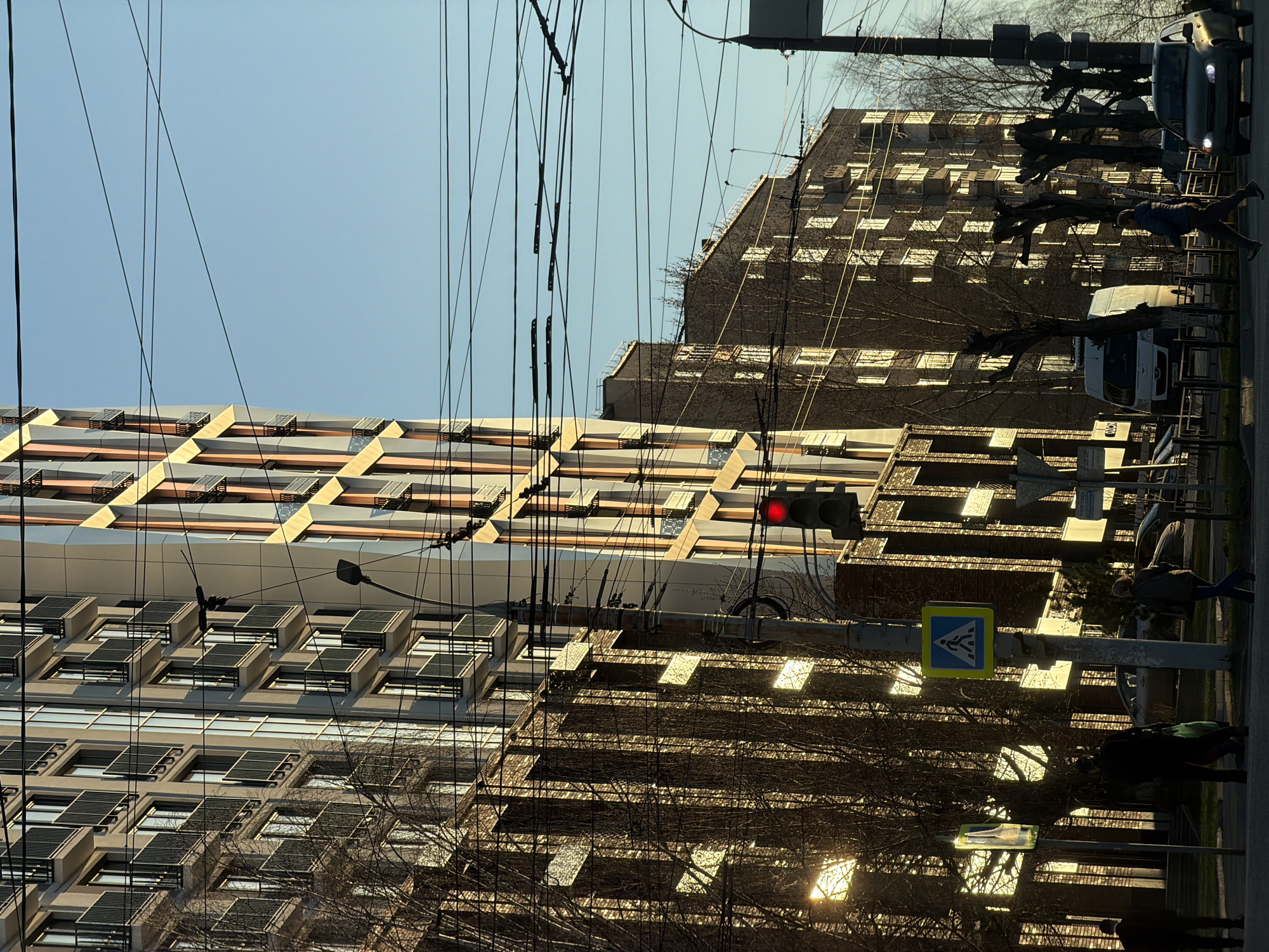 The image, rotated 90 degrees counter-clockwise, depicts a street scene in Izhevsk, Russia. The sky is pale blue. Two multi-story buildings are visible. The building on the left has a modern design with a grid-like facade and balconies. The building on the right is older and constructed with dark brown brick. Power lines and bare tree branches are in the foreground. A traffic light shows red. A pedestrian crossing sign is visible. Pedestrians and vehicles, including a white van, are partially visible on the street. The sun is low in the sky, casting long shadows.