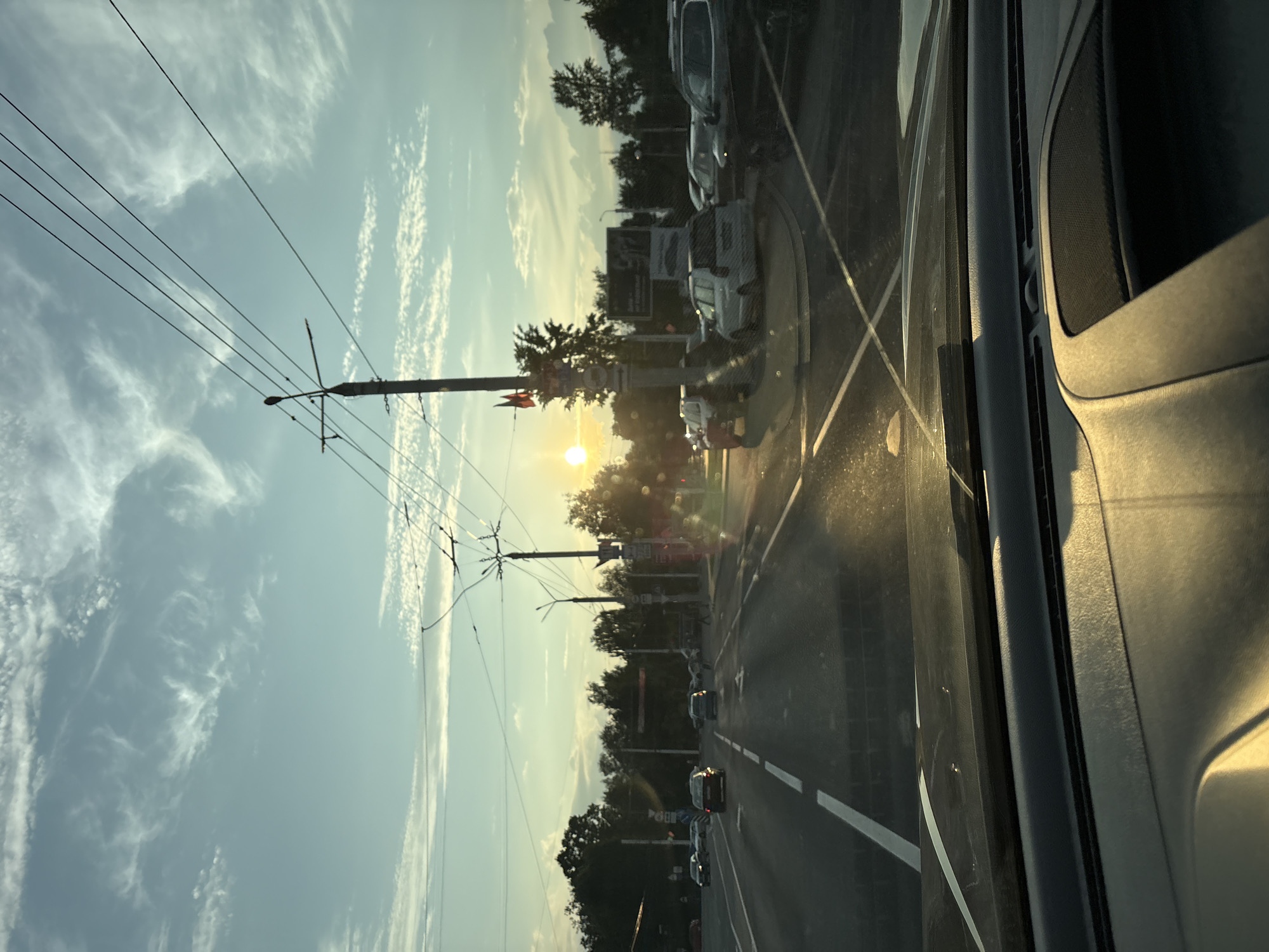 The image depicts a view from inside a vehicle, looking forward through the windshield at a street scene. It is daytime, and the sun is low in the sky, casting a warm, golden light and creating lens flares on the windshield. The sky is a pale blue with wispy white clouds. Power lines and utility poles are prominent in the foreground and midground, crisscrossing the sky.
The street is paved and appears to be a multi-lane road with white lane markings. Several cars are visible on the road, driving in the same direction as the viewer. On the sides of the road, there are trees and buildings, suggesting an urban or suburban environment. Some pedestrian traffic can be seen on the sidewalks.
The location is identified as Vitsebsk, Belarus. The scene appears to be a typical urban street during daylight hours, possibly late afternoon given the low angle of the sun. There are no specific landmarks that can be identified without further information, but the presence of overhead power lines and the general architectural style of the visible structures are consistent with urban environments in Eastern Europe.
The overall impression is one of a mundane, everyday scene viewed from a car, with the golden hour light adding a touch of visual interest.