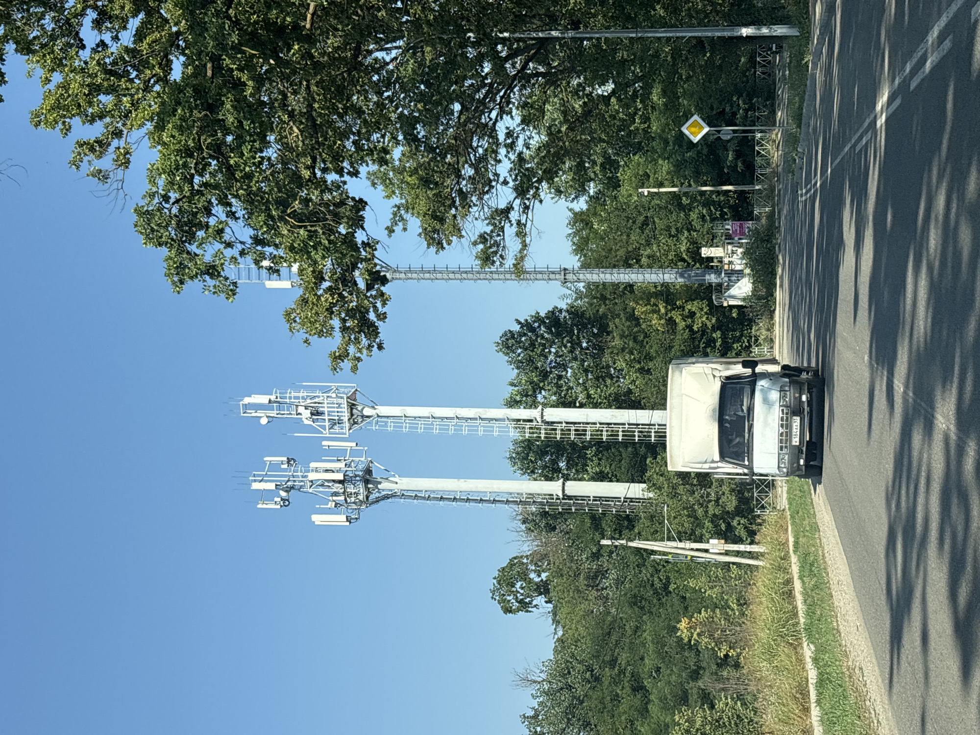 This is a photograph of a road in the city of Yuzhny, Russia, on a clear, sunny day. In the foreground, a white truck with a large cargo box is driving down the asphalt road. The truck has a license plate that reads "K184AA 33". To the left of the road, there is a grassy shoulder and a dense line of green trees. In the background, three tall, silver telecommunications towers stand against a bright blue sky. These towers are equipped with numerous antennas and access ladders. The trees continue behind the towers, creating a lush, green backdrop. To the right of the road, another telecommunications tower is visible, partially obscured by the large, leafy branches of an oak tree. A yellow diamond-shaped road sign with a black border is also present on the right side of the road. The overall scene depicts a rural or suburban roadside environment with significant telecommunications infrastructure.